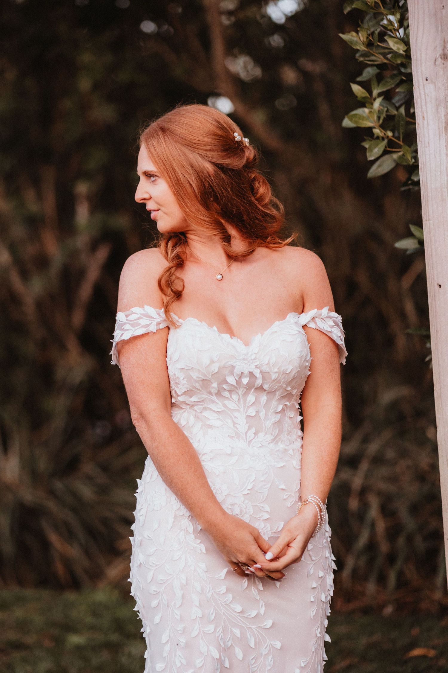 Elegant bride in off-shoulder floral wedding gown poses in garden setting.