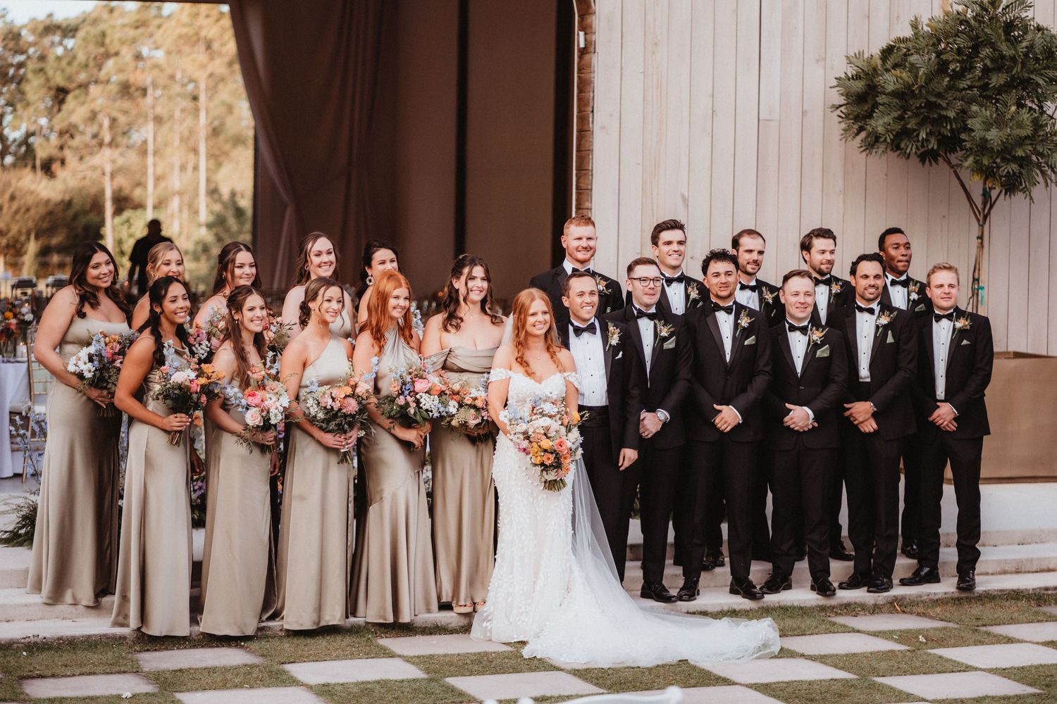 Large wedding party poses formally outside barn venue, bridesmaids in taupe gowns and groomsmen in black tuxedos.