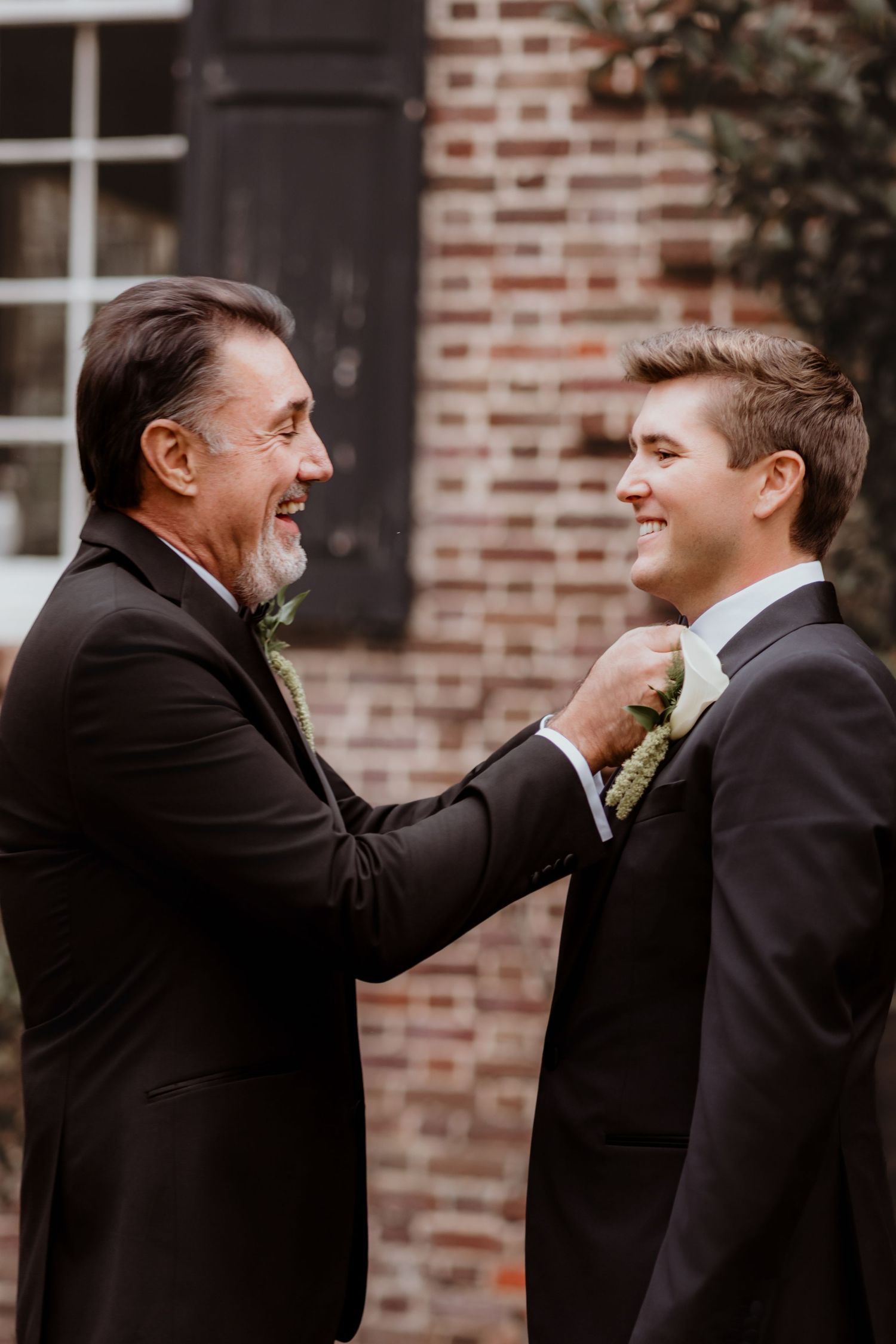 Older man smiling while adjusting boutonniere on laughing groom in black tuxedo against brick wall.