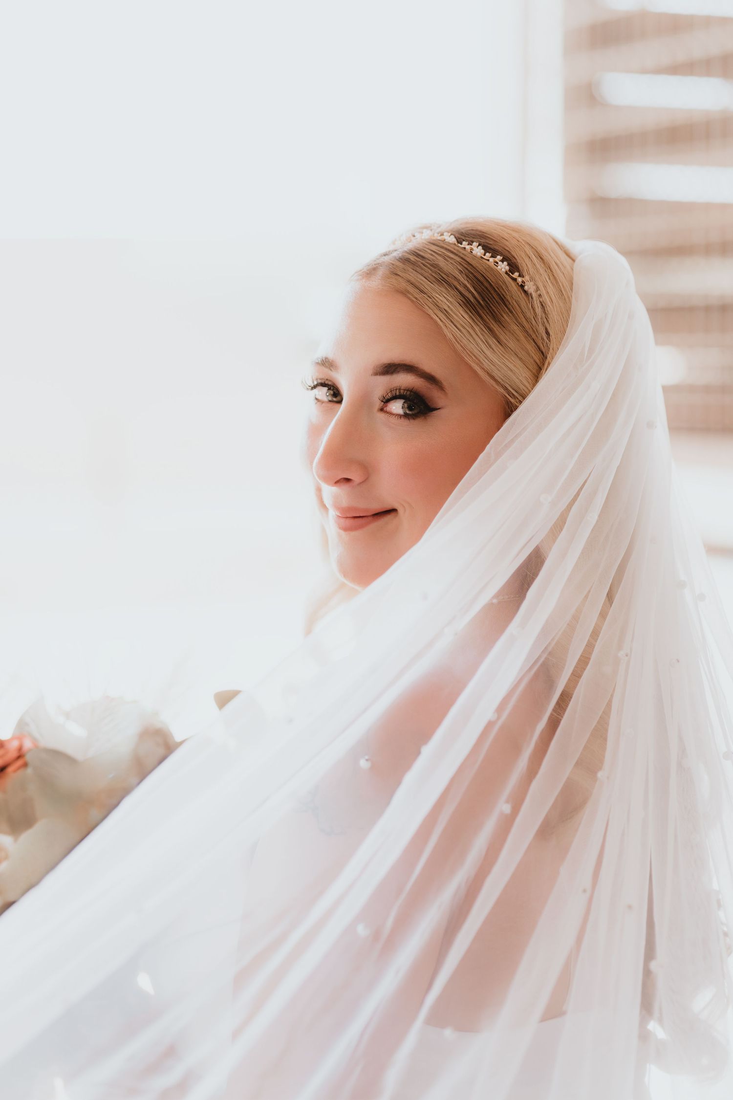 Elegant bride smiles softly wearing a flowing white veil and delicate pearl tiara accessory.
