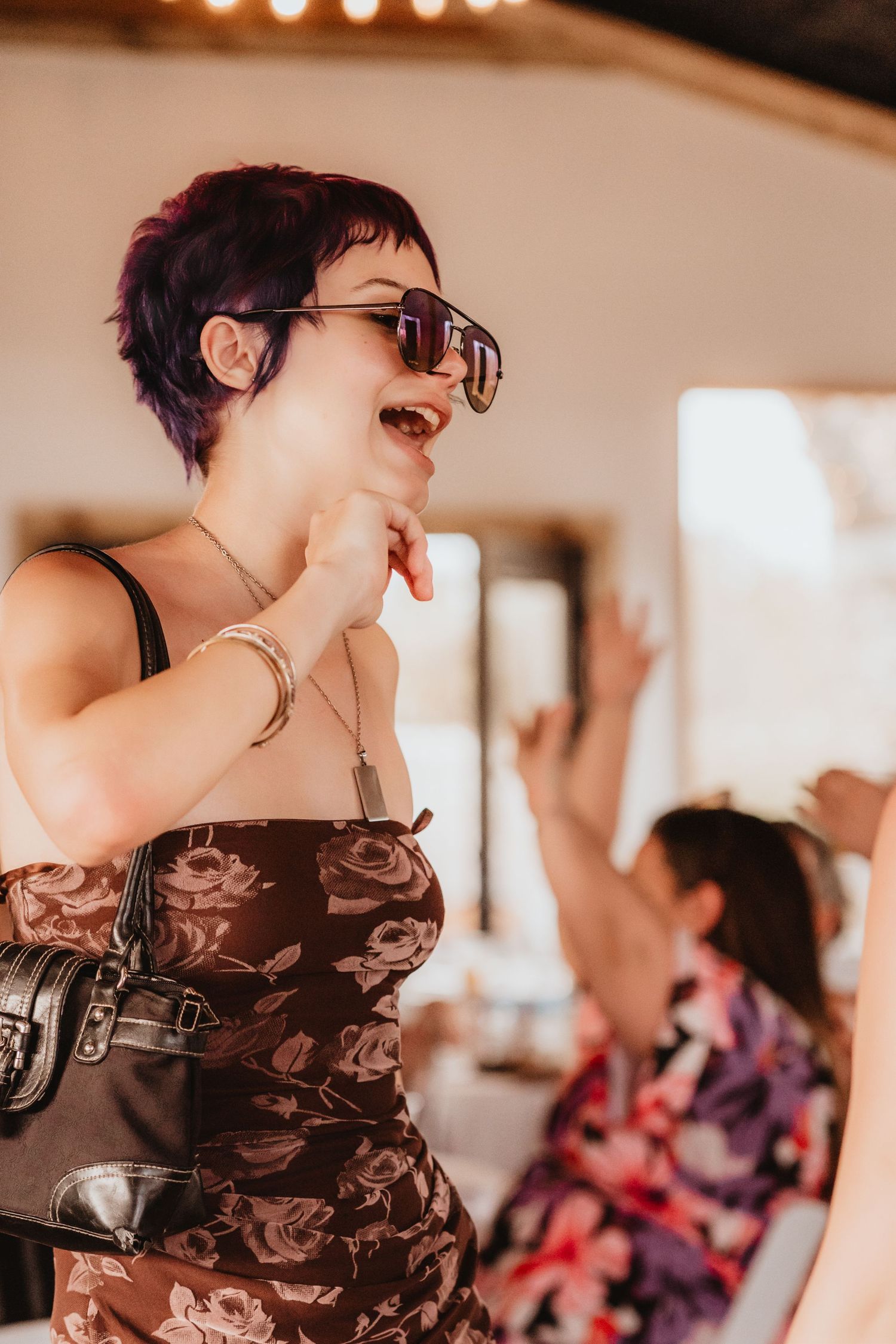Woman with purple pixie cut and sunglasses laughs while dancing at a wedding reception.