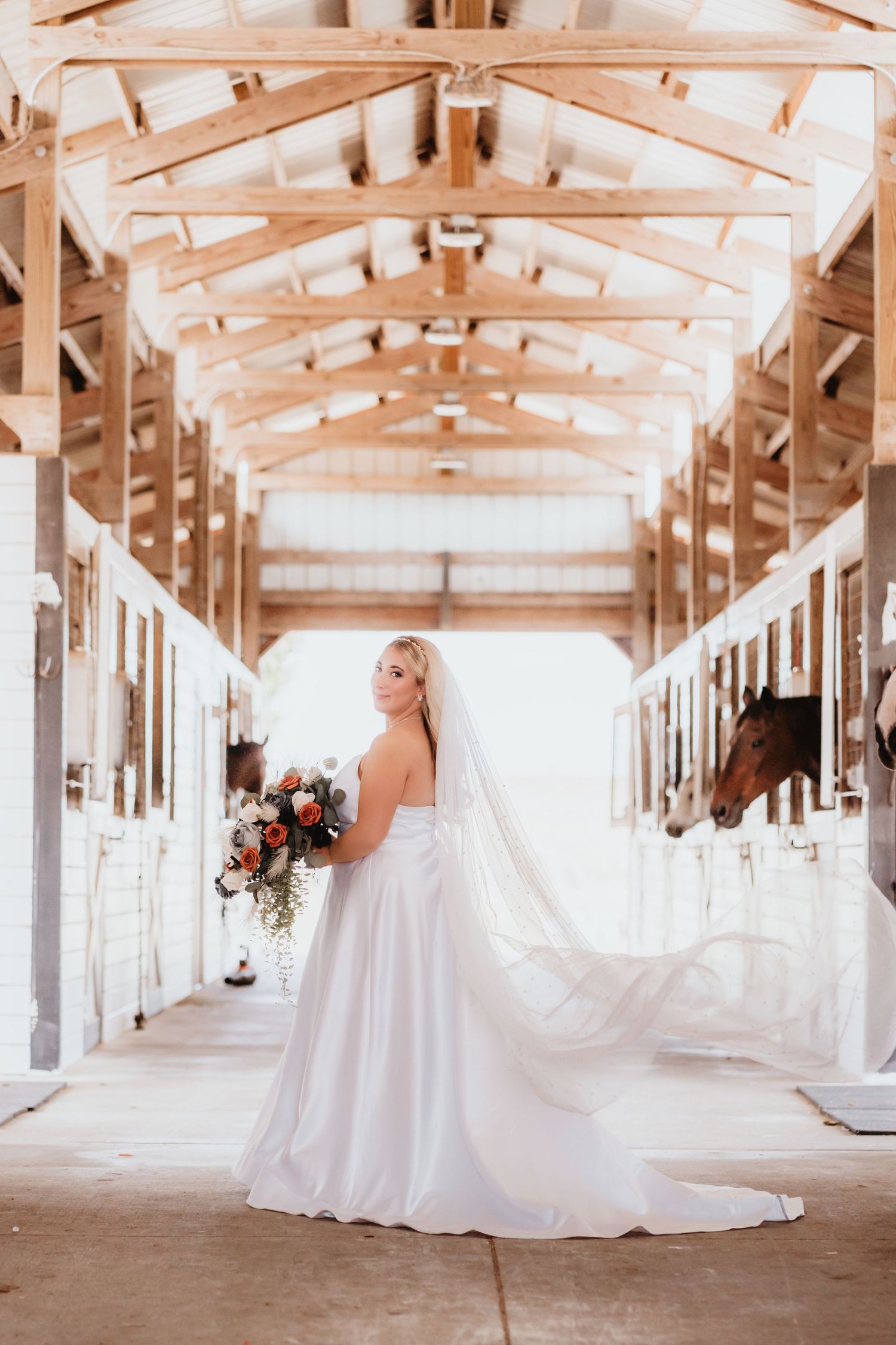 Bride in a white gown with a flowing veil poses in a horse stable holding an orange and gray bouquet.