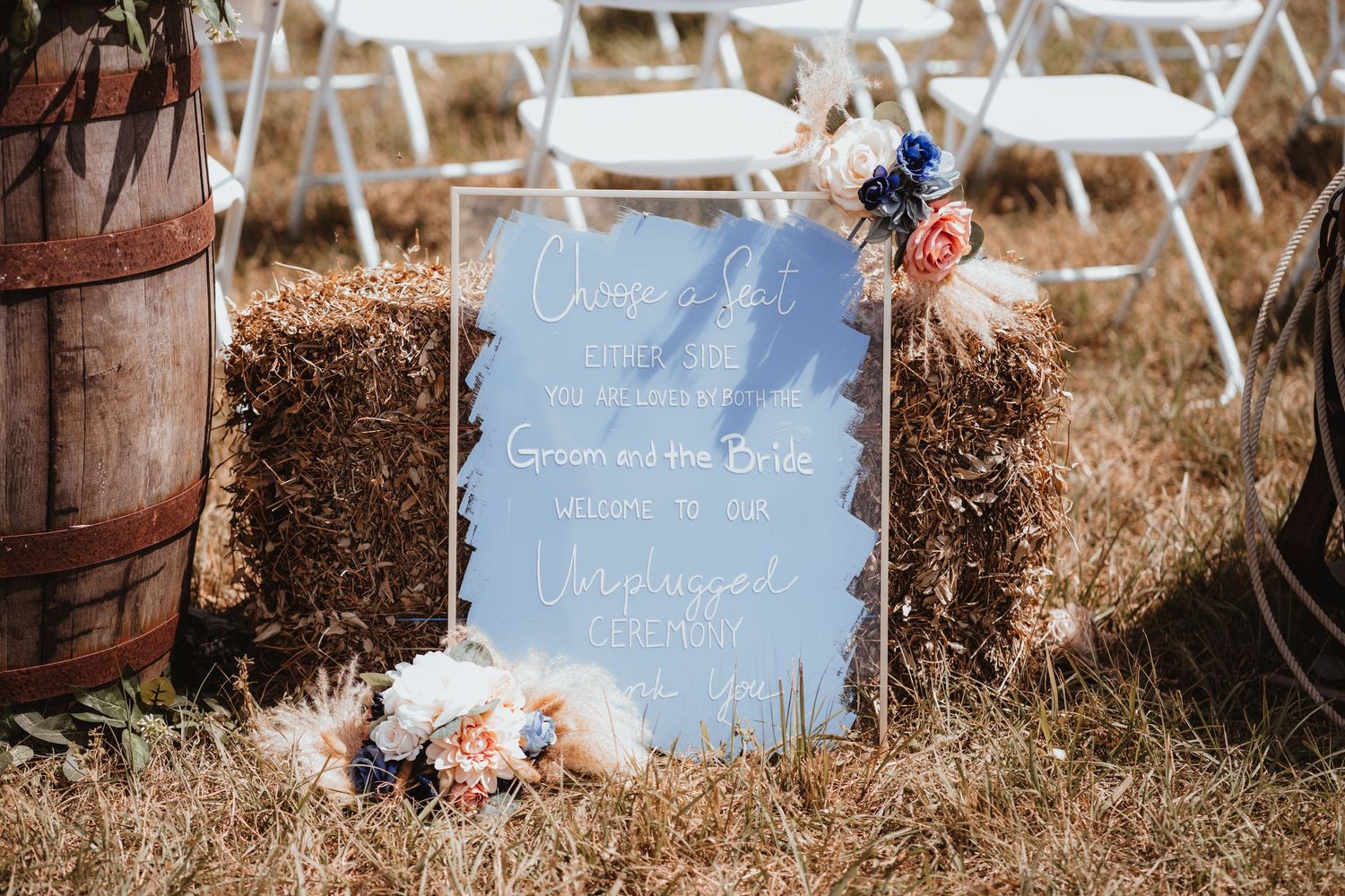 Blue wedding welcome sign on hay bale with floral accents reading Groom and Bride welcome message at rustic outdoor ceremony.