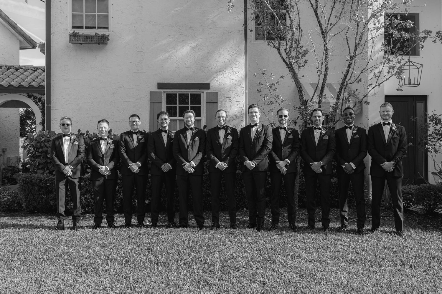 Black and white photo of groom and groomsmen lined up in tuxedos outside venue.