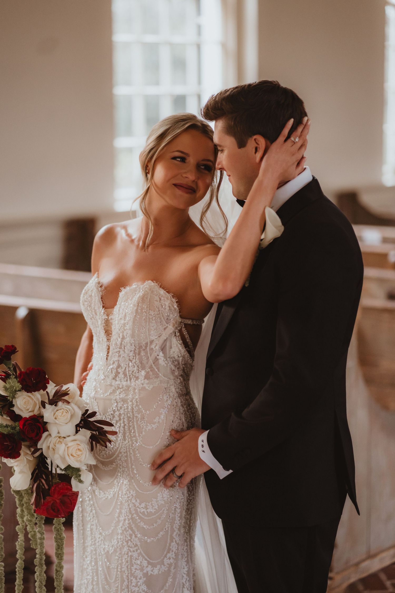 Smiling bride in beaded gown tenderly holds grooms face in a sunlit church interior.