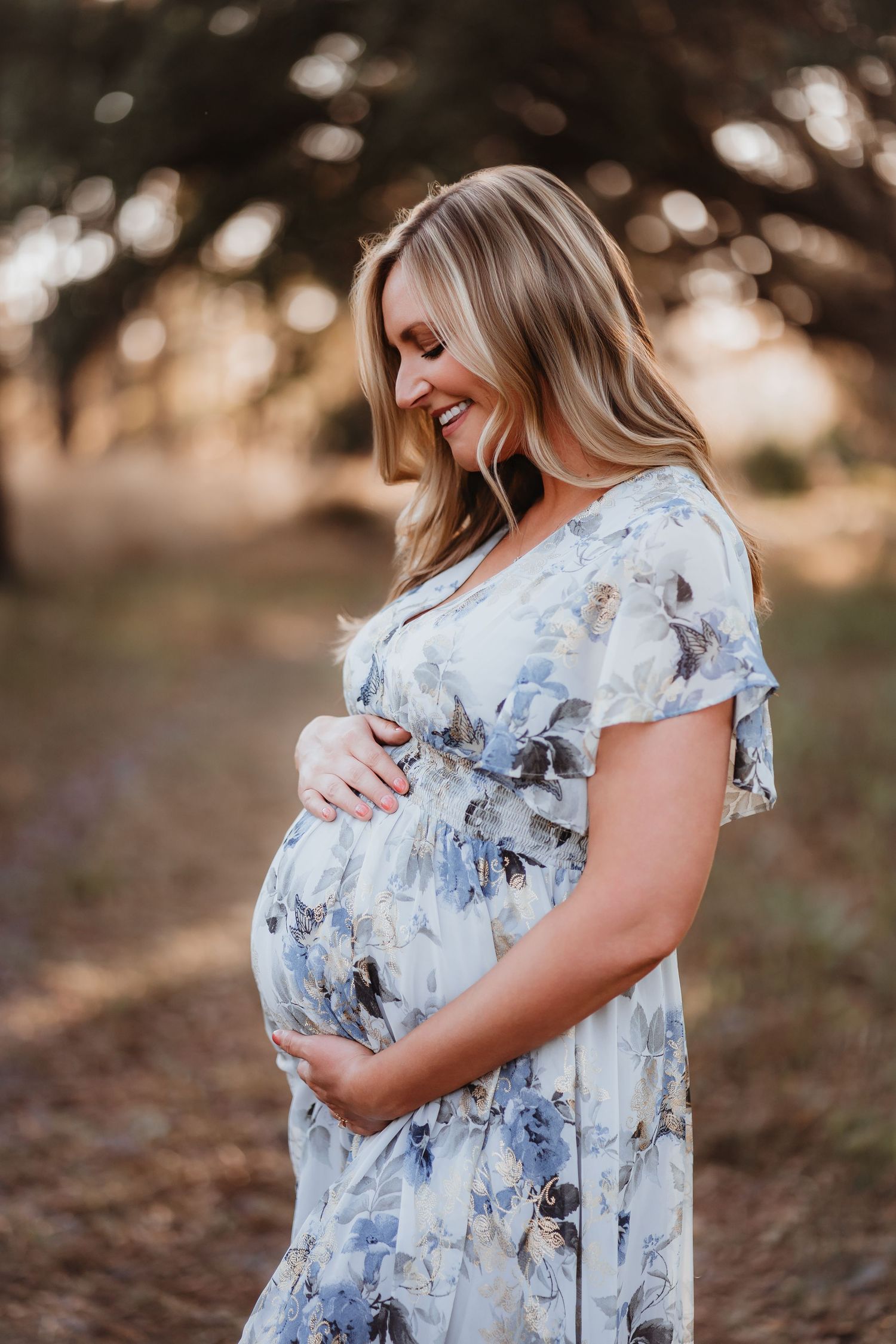 Smiling pregnant woman in floral dress cradling baby bump outdoors in golden light.