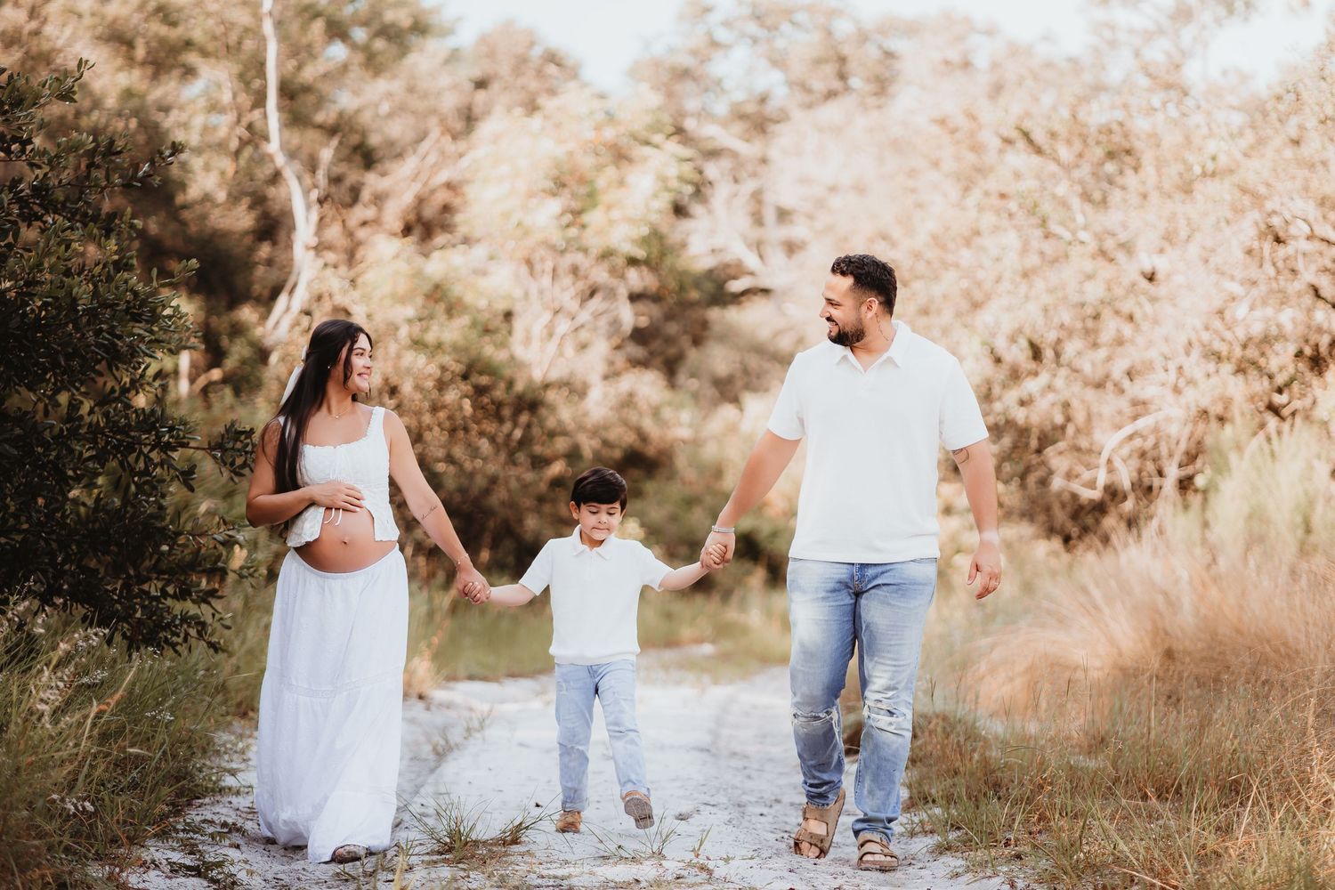 Family maternity shoot with pregnant mother, father, and child holding hands on sunny nature path.
