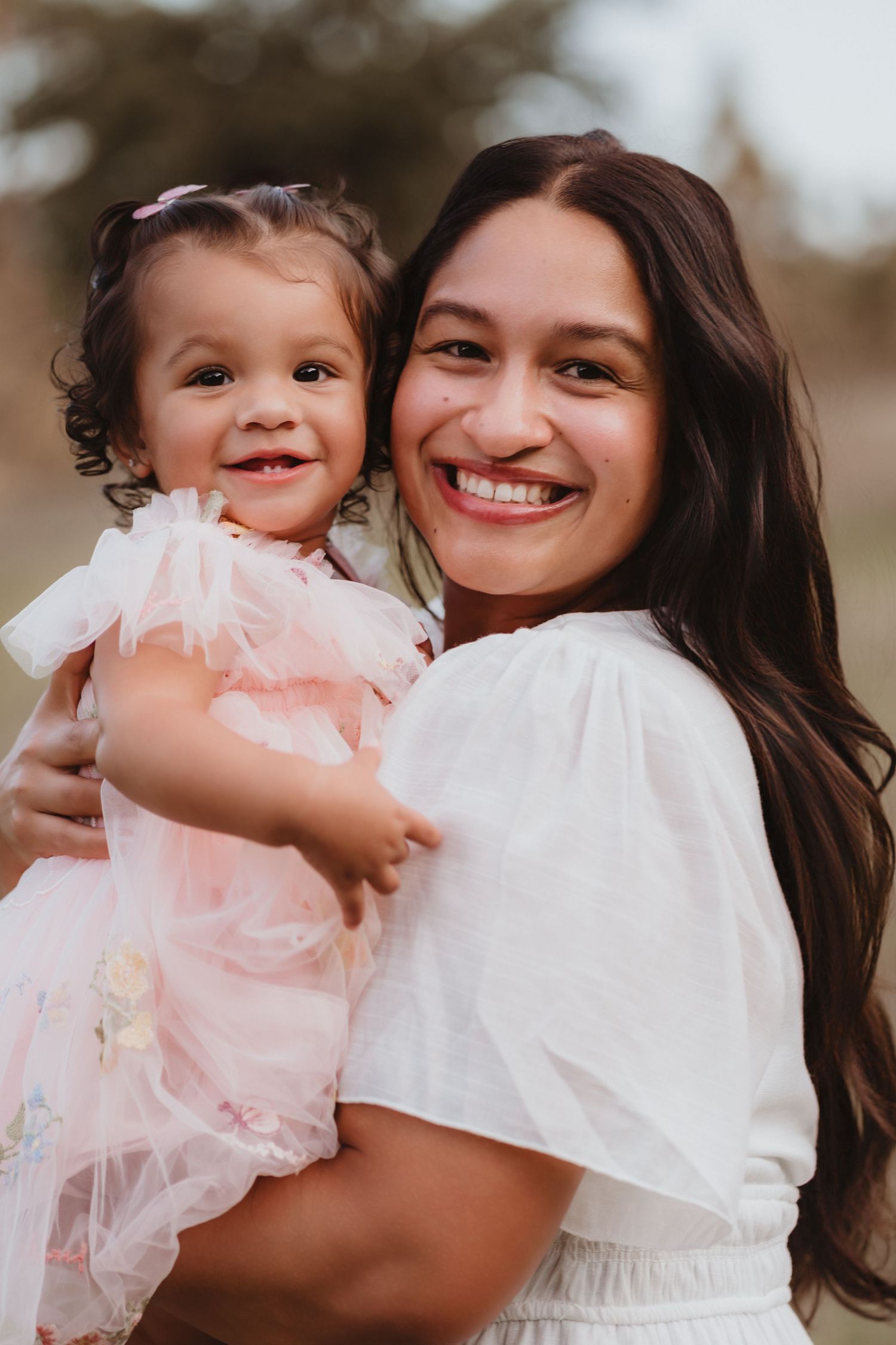 Smiling mother in white dress holding toddler girl in pink tulle dress outdoors.