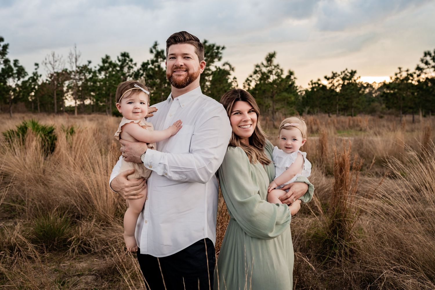 Family of four with two babies posing in golden field at sunset, parents in white and green.