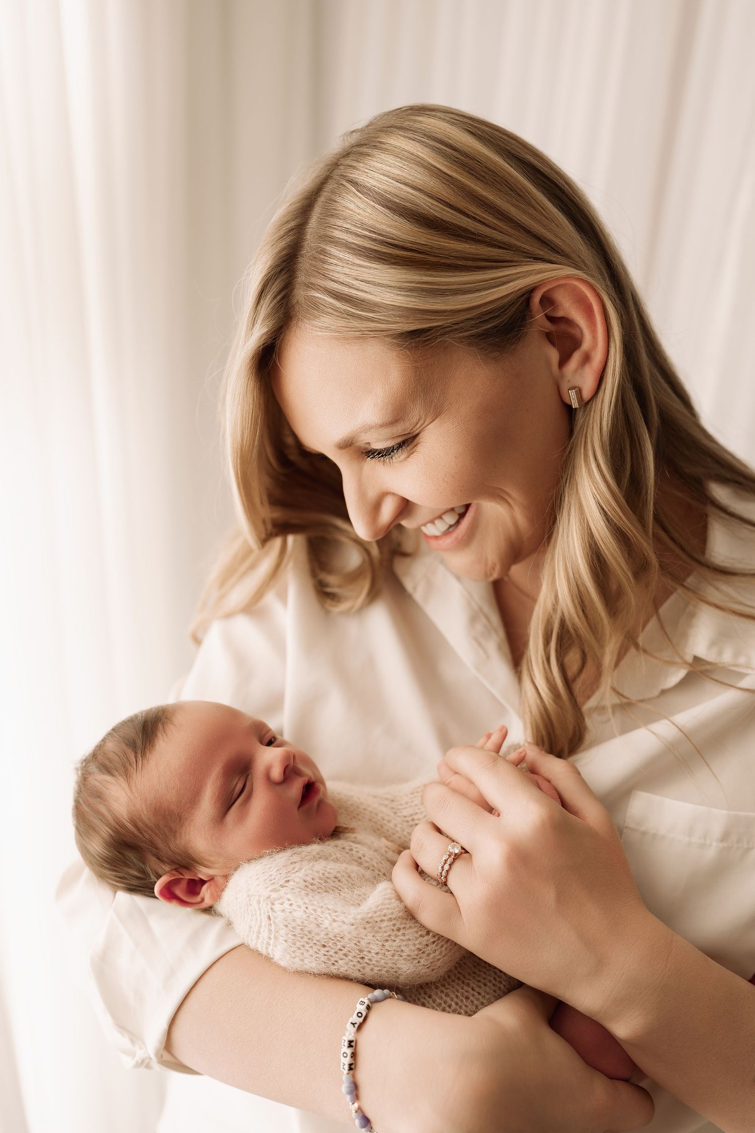 Smiling blonde mother looking lovingly at her sleeping newborn dressed in a beige knit outfit.