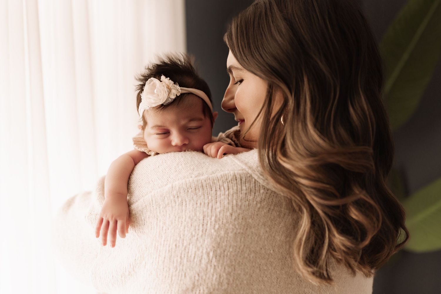 Mother in cream knit sweater lovingly holding sleeping newborn with floral headband near window.