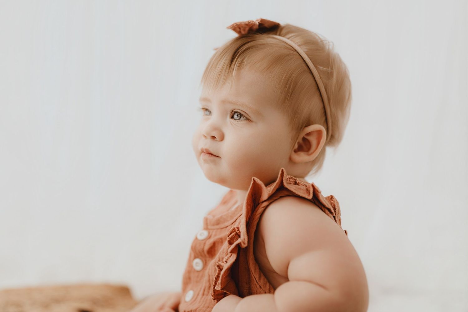 Adorable baby girl in rust ruffle romper with bow headband looking up softly.