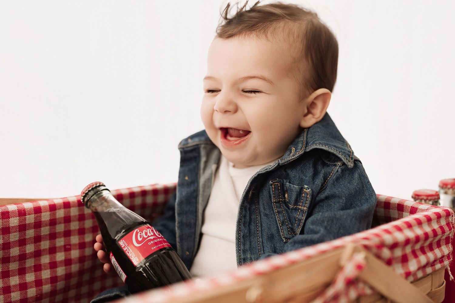 Joyful baby boy in denim jacket laughing while holding a Coca-Cola glass bottle.