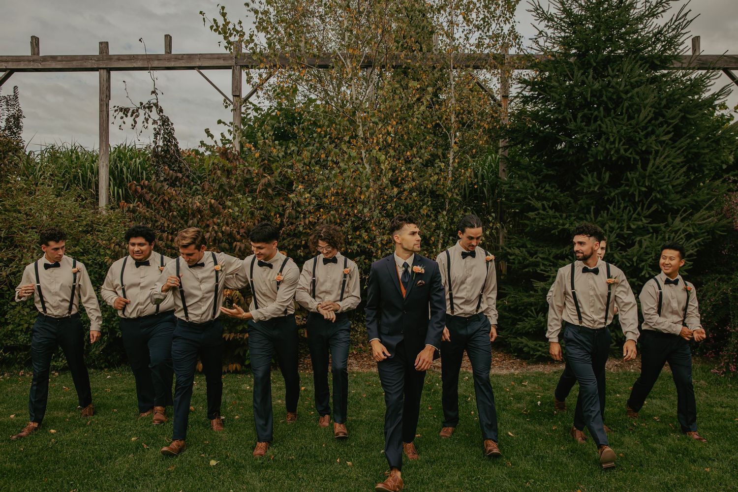 Groomsmen in suspenders and navy attire pose outdoors with wooden pergola backdrop.