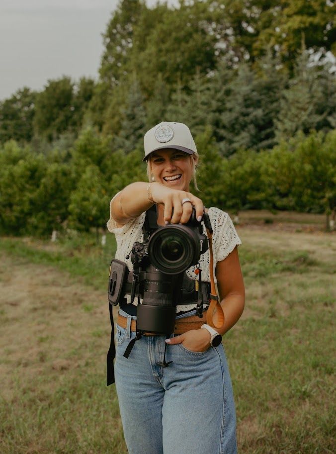 Smiling woman holding a DSLR camera outdoors in a green field, wearing a gray cap and jeans.