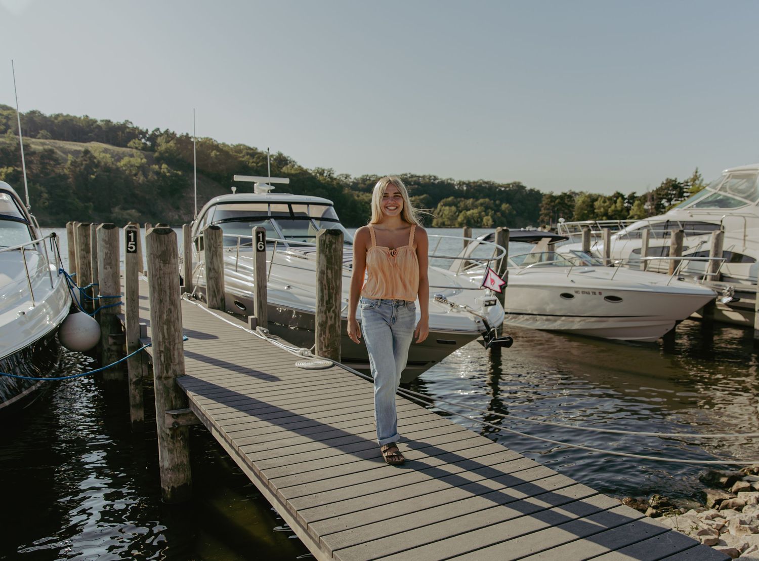 Wooden dock extends over calm water with boats and hills in the background.