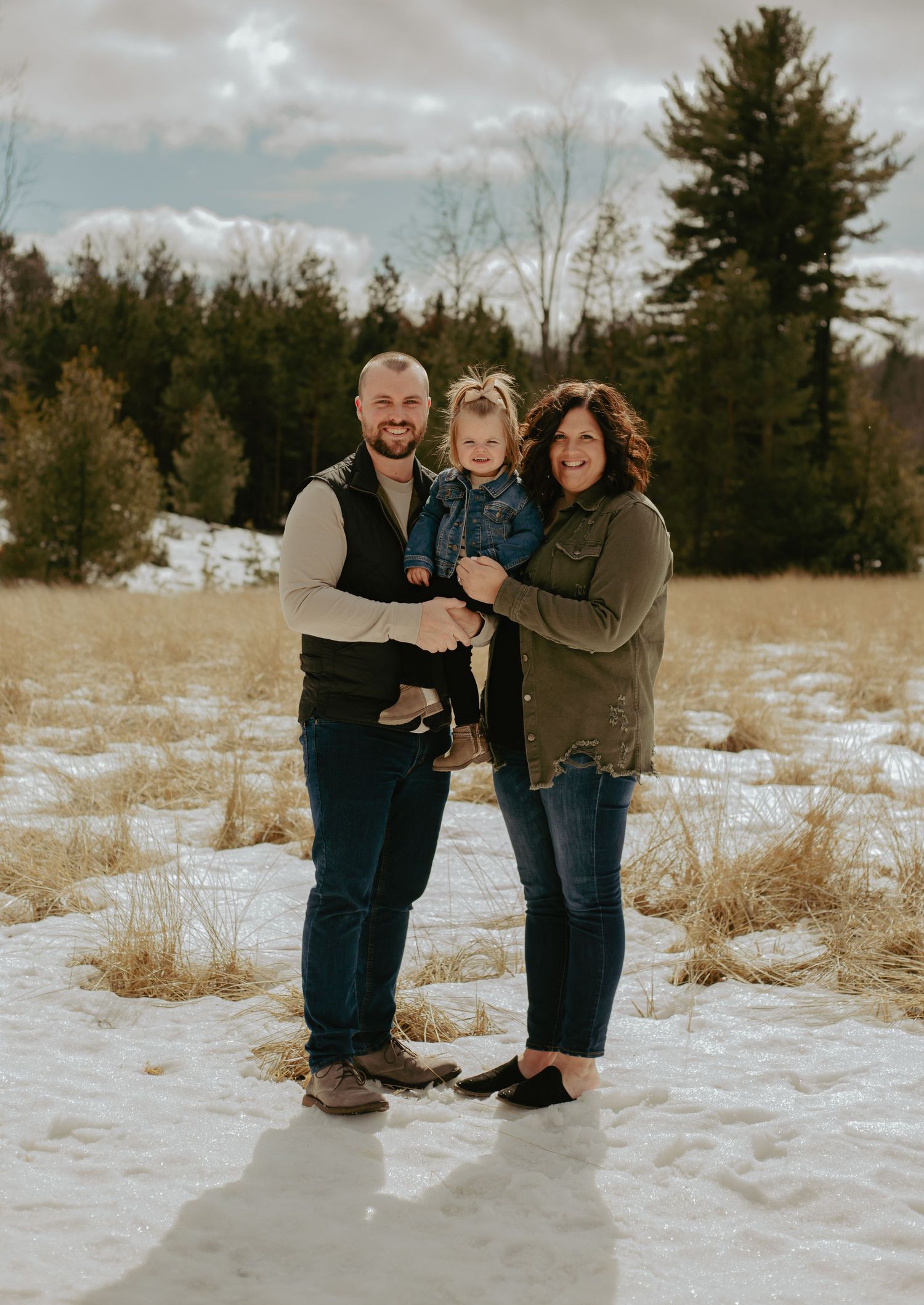 Family enjoying a winter photo session together in a snowy field with evergreen trees in the background.