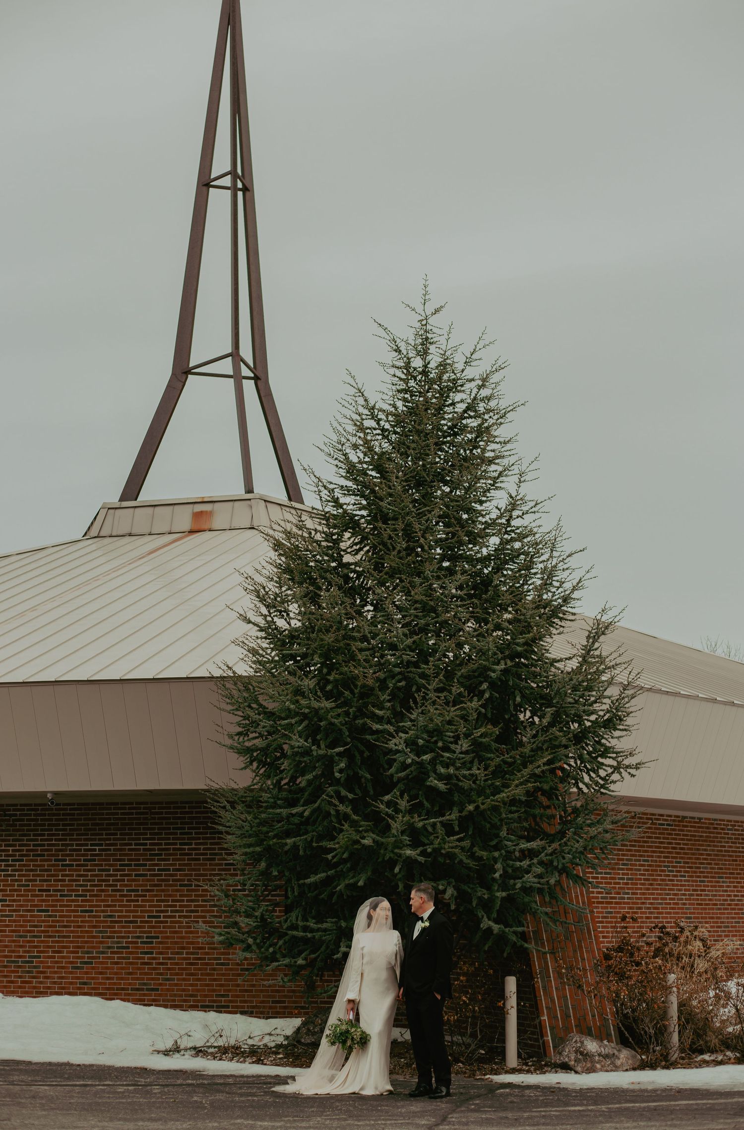 Wedding couple poses near a church with evergreen trees and steeple in background.