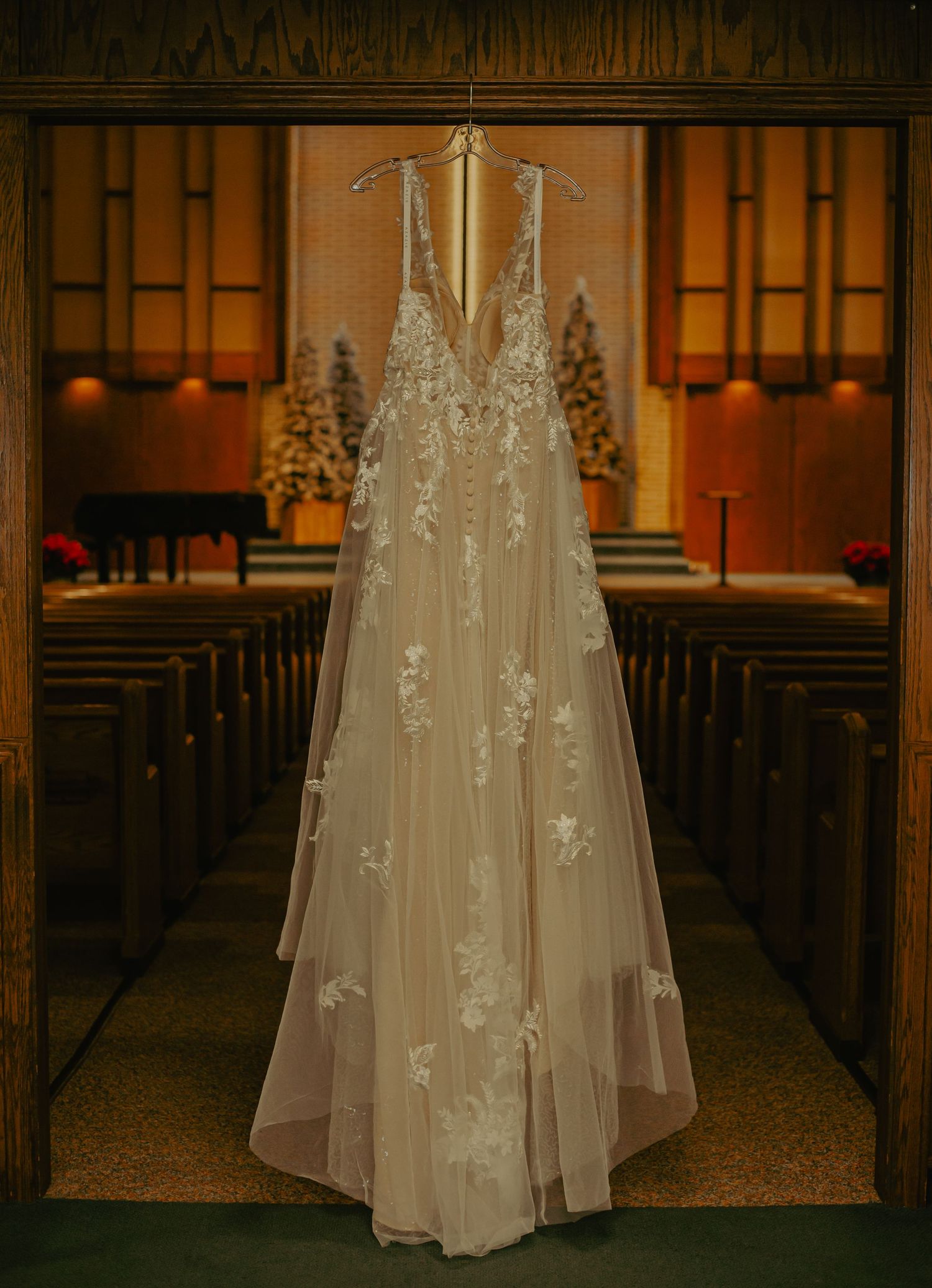 Elegant lace wedding dress hanging in church aisle with warm lighting and wooden pews.