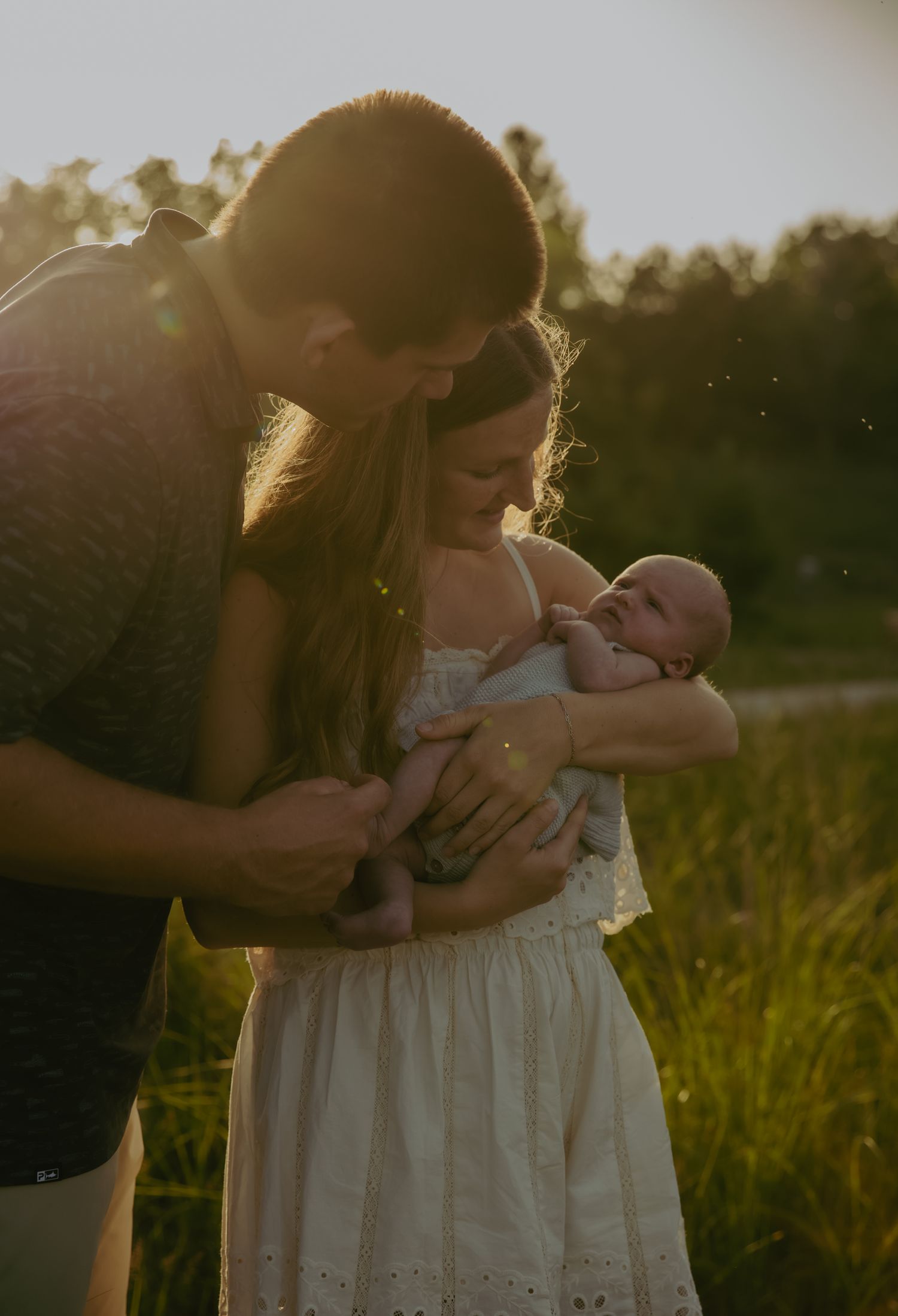 Couple gazing lovingly at newborn baby outdoors in golden sunset light surrounded by lush green grass.