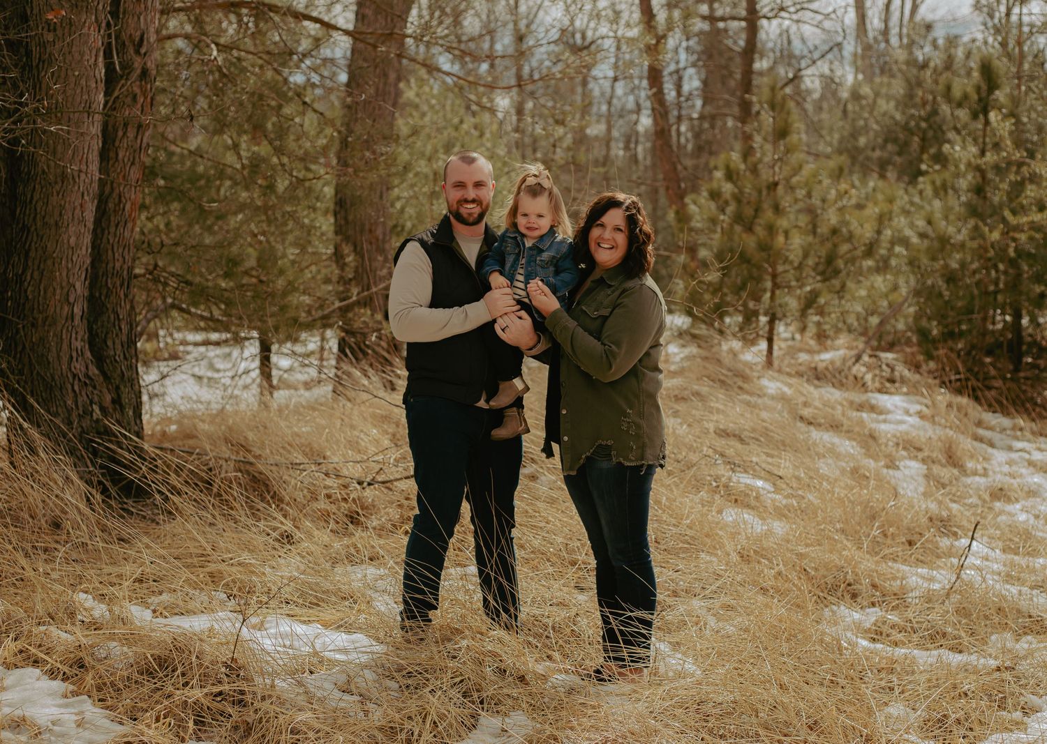 Family photo session in a wooded area with snow patches on the ground.