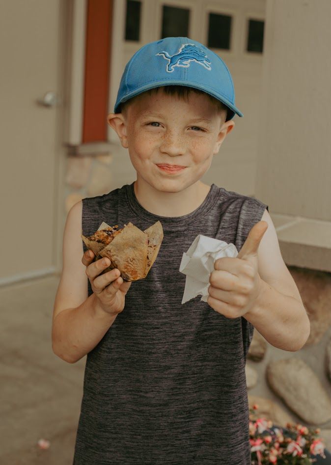 A smiling boy in a blue Detroit Lions cap gives a thumbs up while holding a taco outdoors.