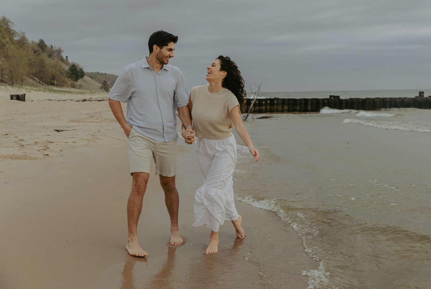 Happy couple walks hand-in-hand along sandy beach shoreline, dressed in casual neutral tones on cloudy day.