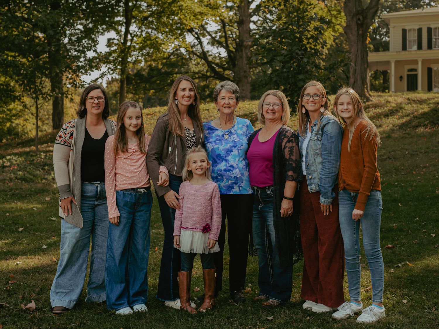 Multi-generational family group of women and girls smiling outdoors on green lawn with trees and historic home.