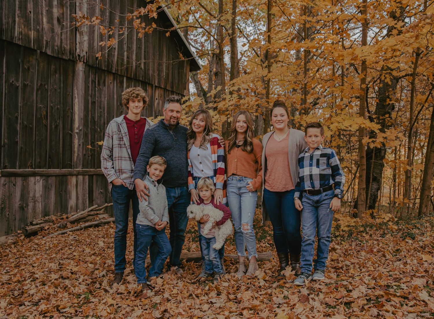 Family photo session in autumn forest with golden leaves and rustic barn backdrop.