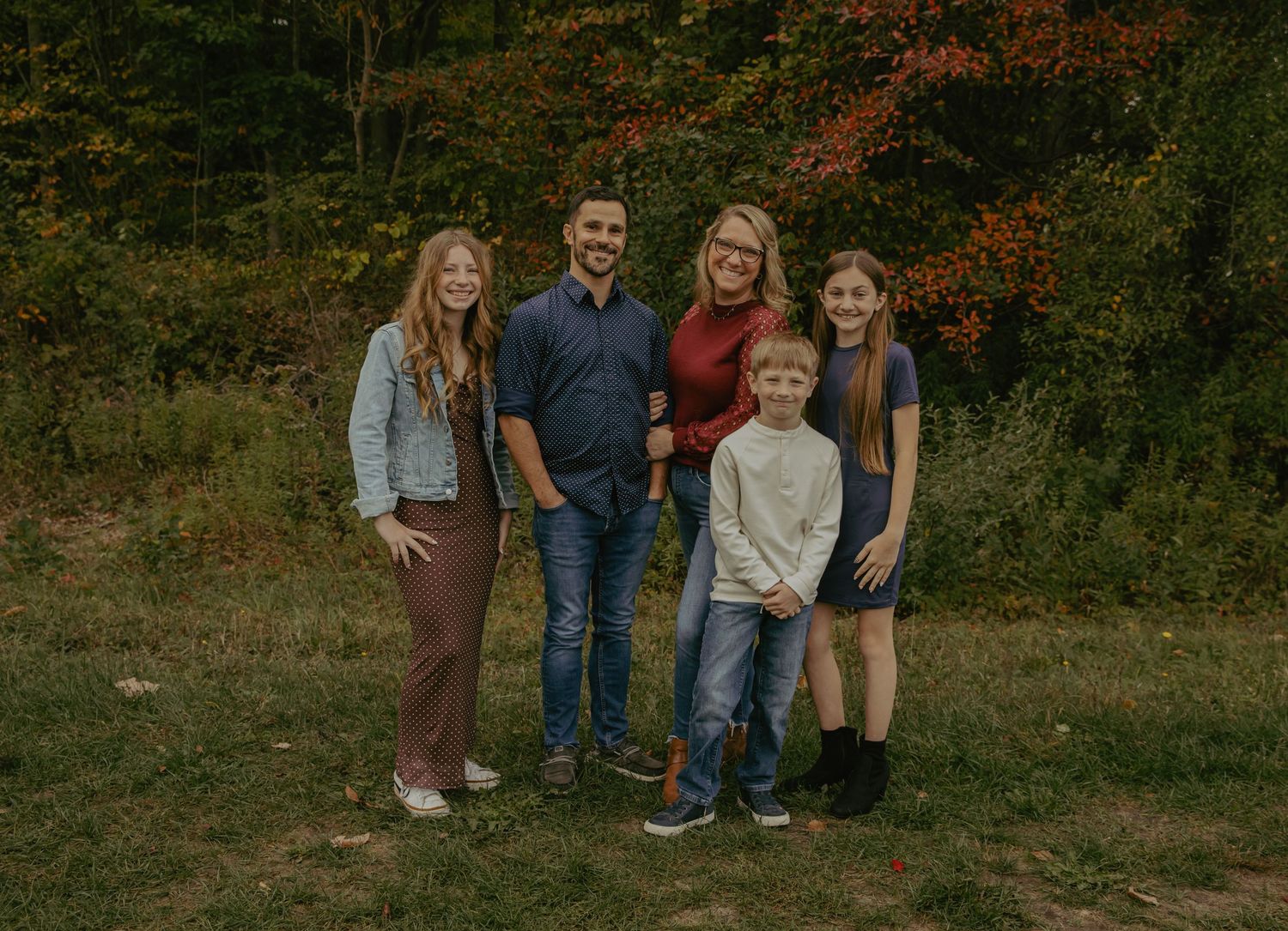 Family of five smiling outdoors in autumn, surrounded by colorful fall foliage in a scenic forest setting.
