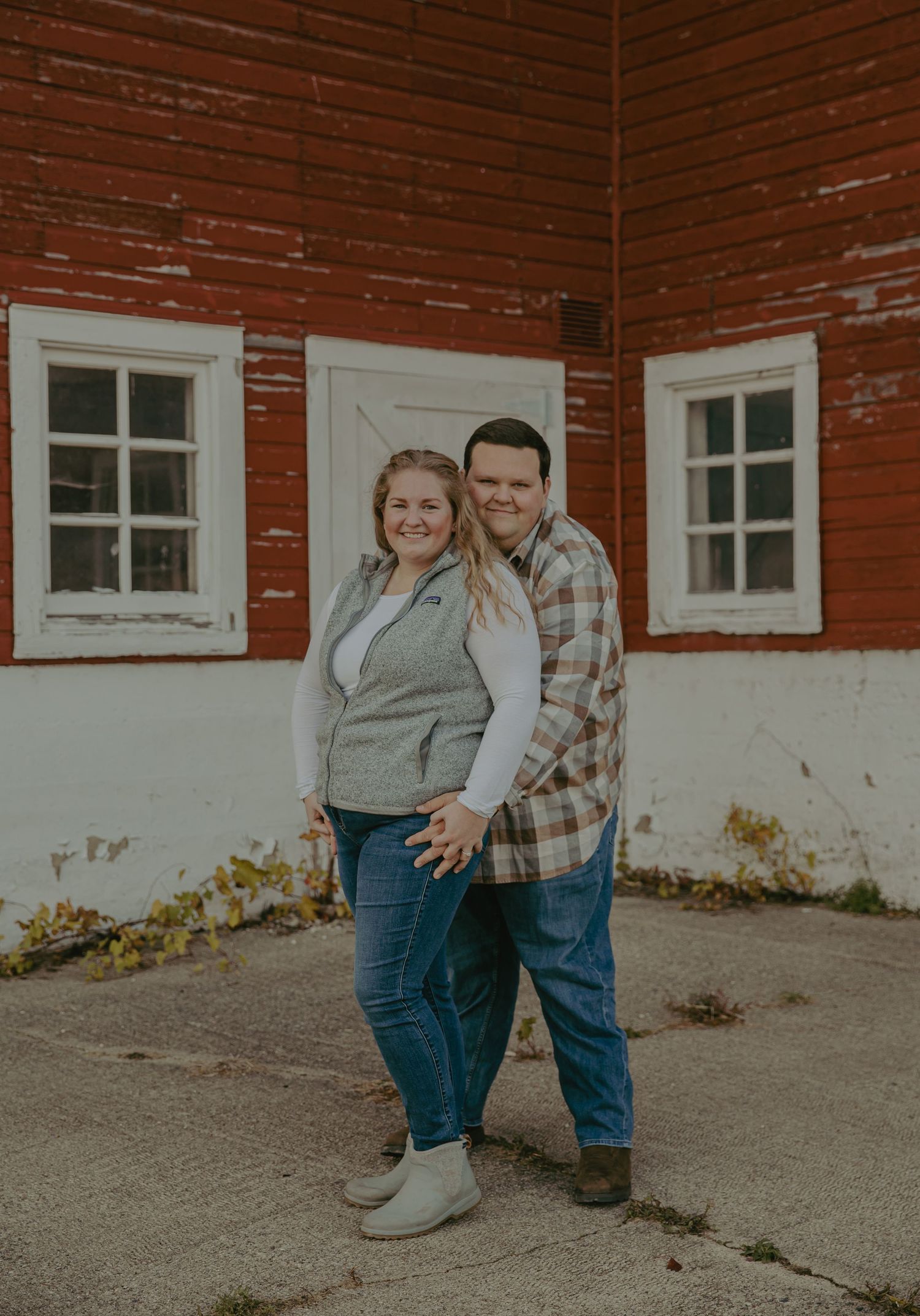 Couple posing together against red barn wall with white windows.