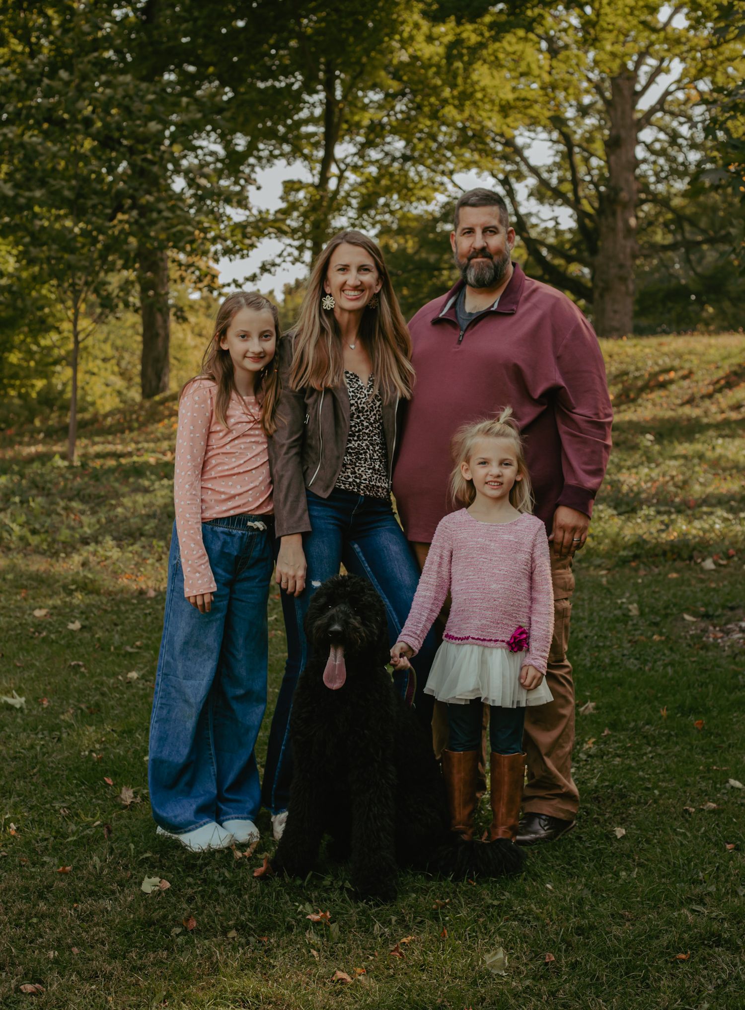 A family poses together outdoors in casual clothing against a backdrop of autumn trees.