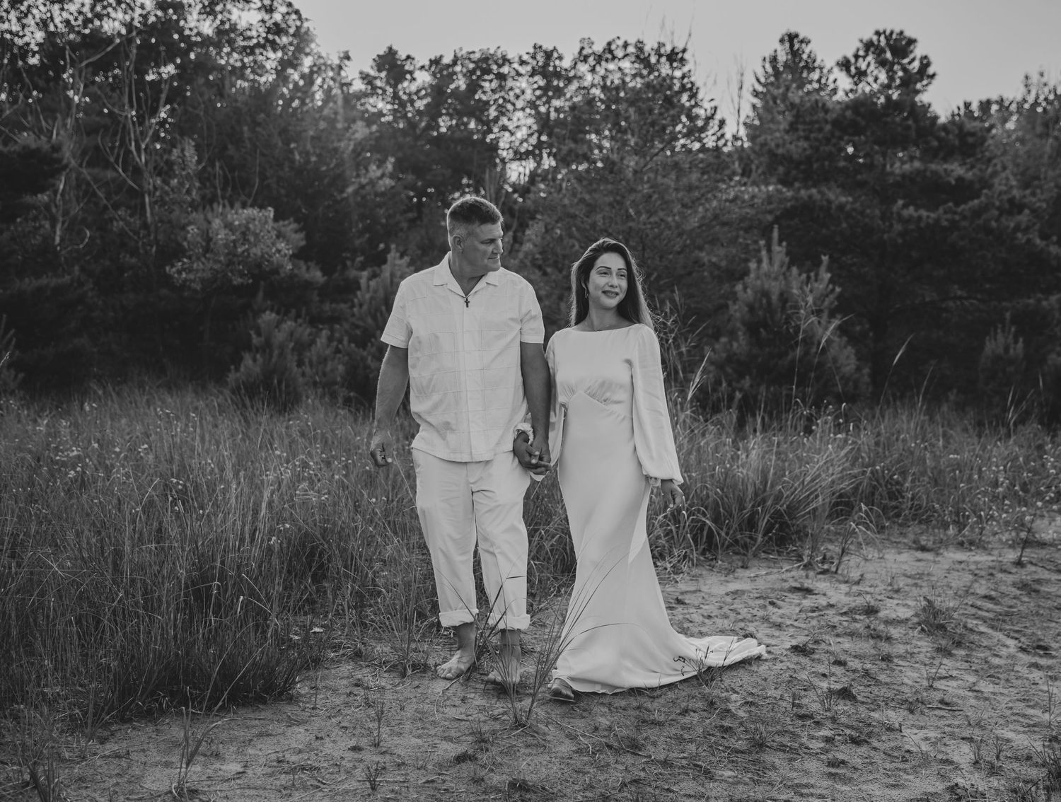 Couple in white attire walking together on sandy beach path.
