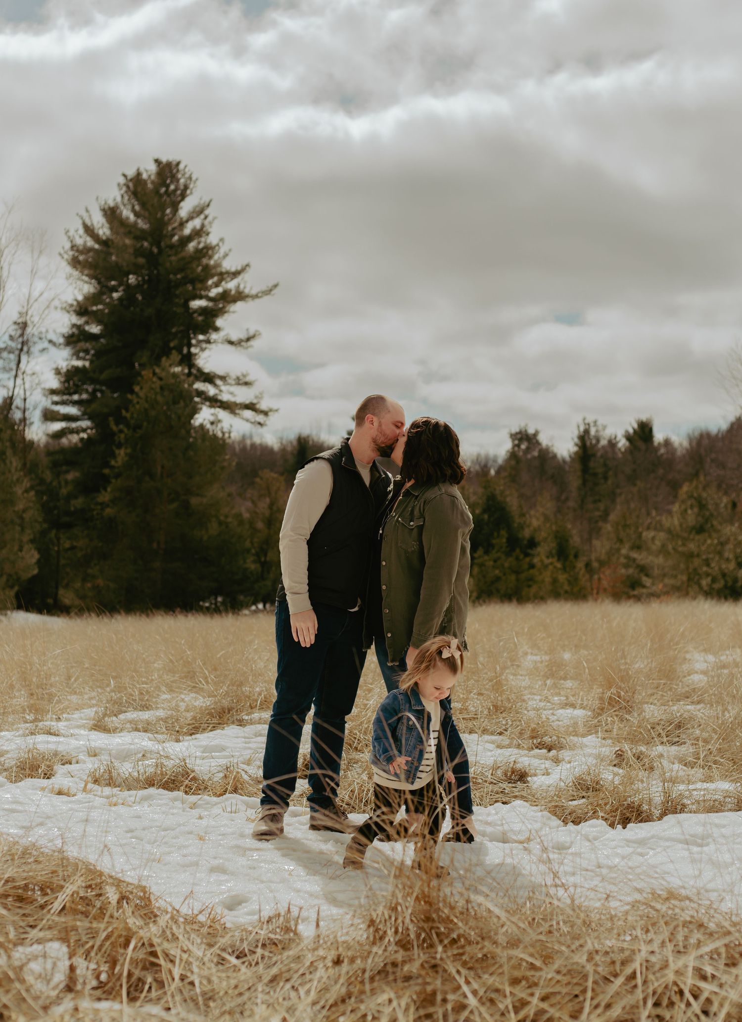 A couple strolls through a snowy winter field with tall dried grass and evergreen trees.