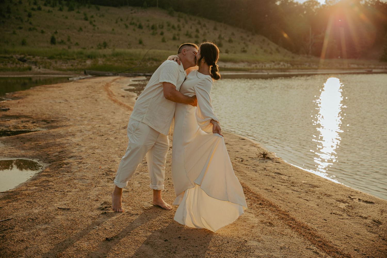 Newlyweds dancing on beach at sunset with water reflection.