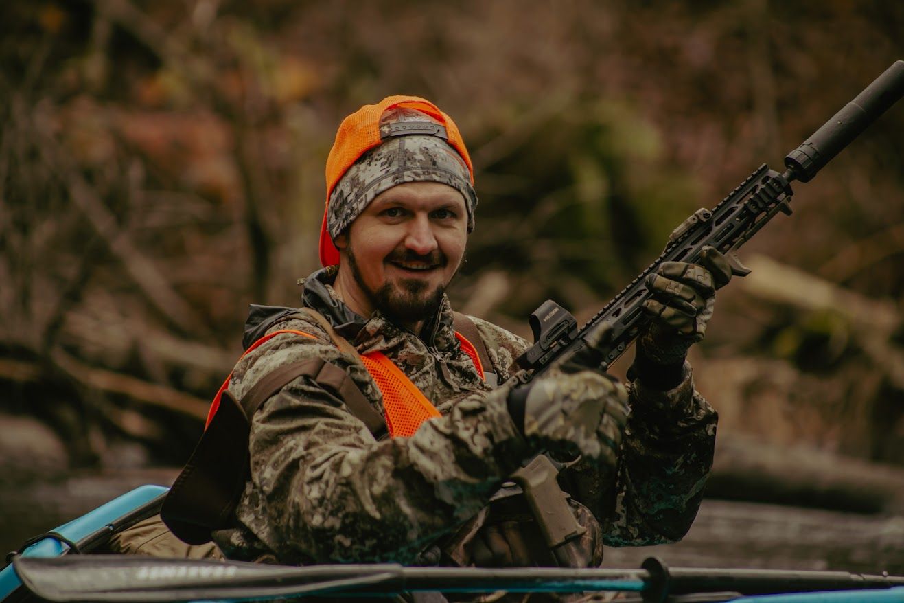 A hunter in camouflage gear and orange beanie holds a rifle while sitting in a boat during hunting season.