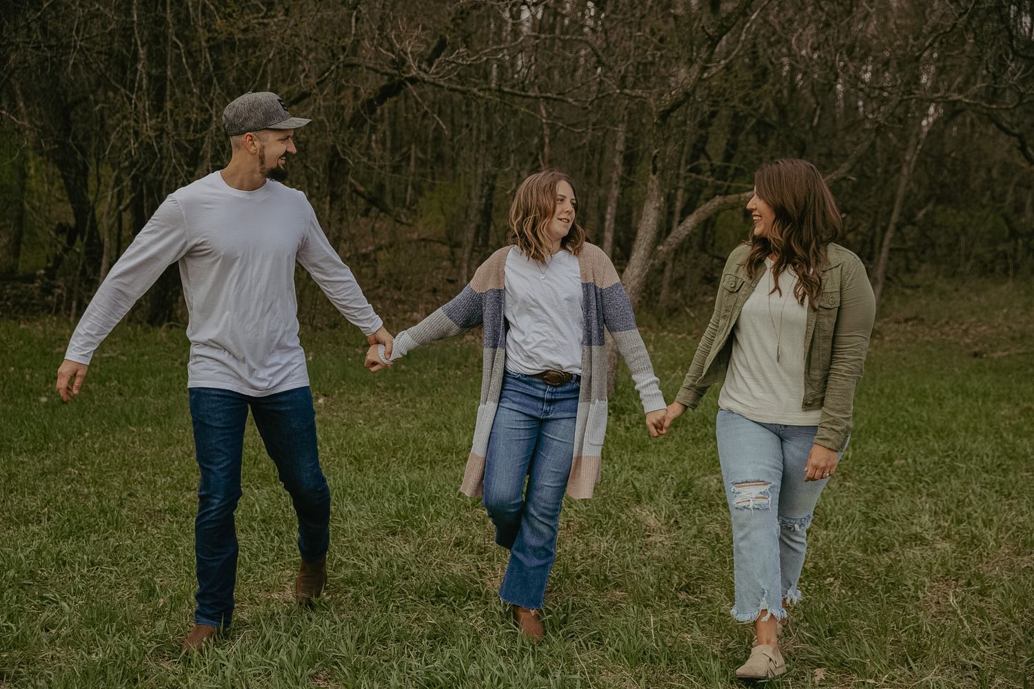 Three friends hold hands while walking through a lush green meadow surrounded by trees in casual fall outfits.