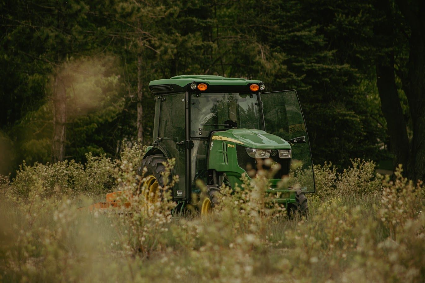 Green tractor visible through tall grass and wildflowers in a rural field.