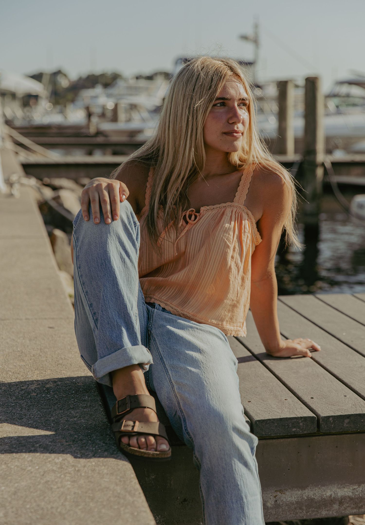 Woman in overalls sitting on dock with boats in background.