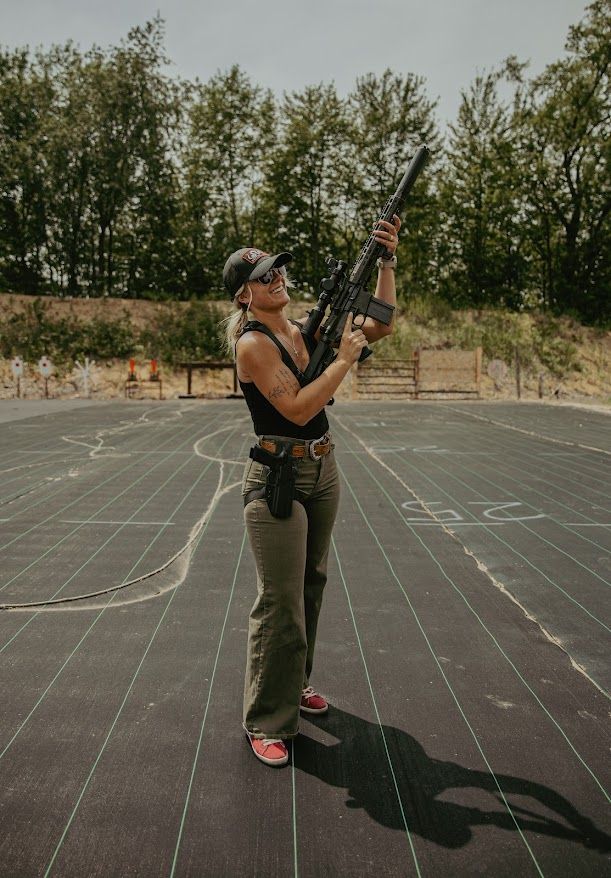 Person in tactical gear holding a camera on a basketball court with white lines and trees in the background.