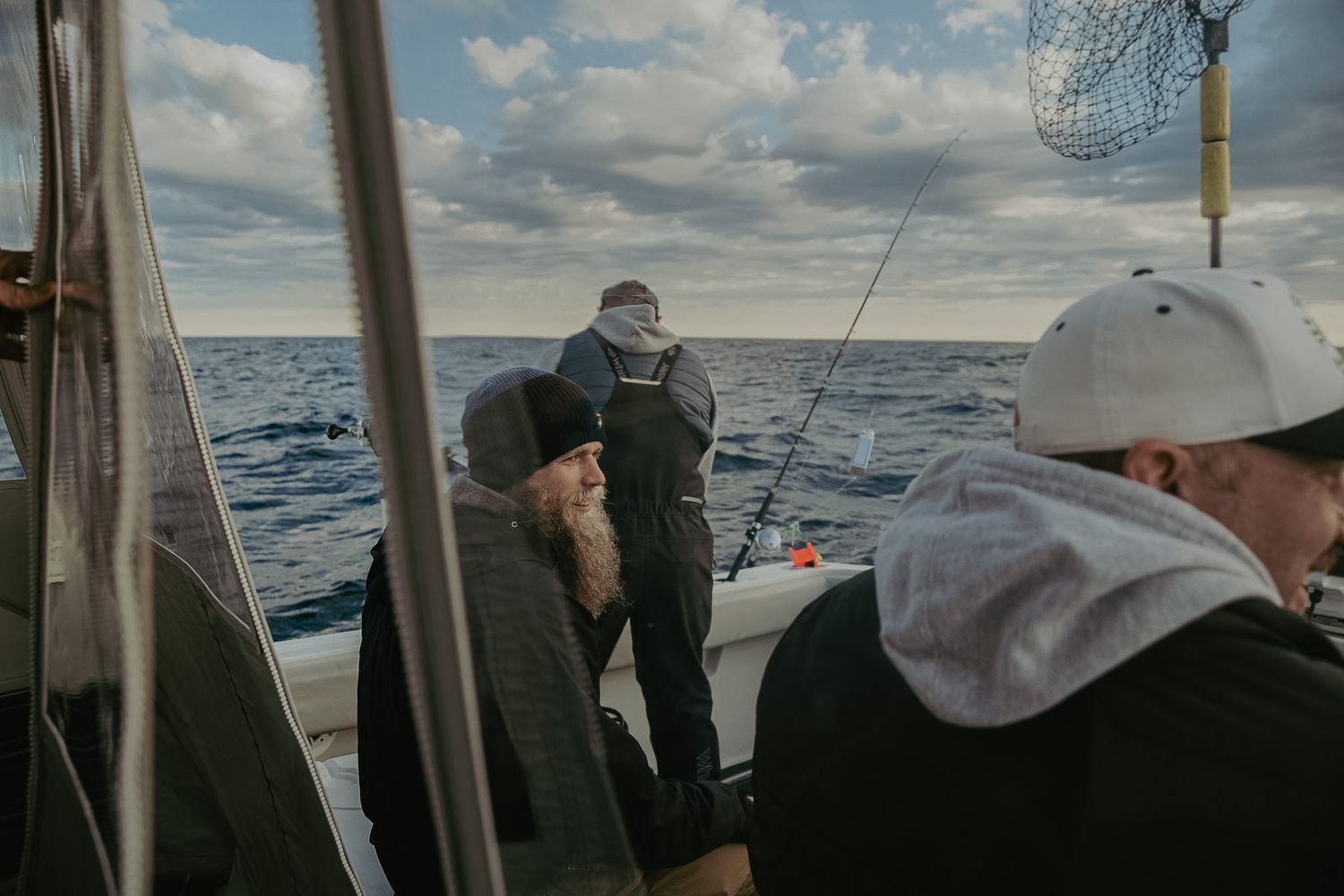 Men fishing on a boat in open ocean waters, with fishing rods and a net visible against a cloudy sky.