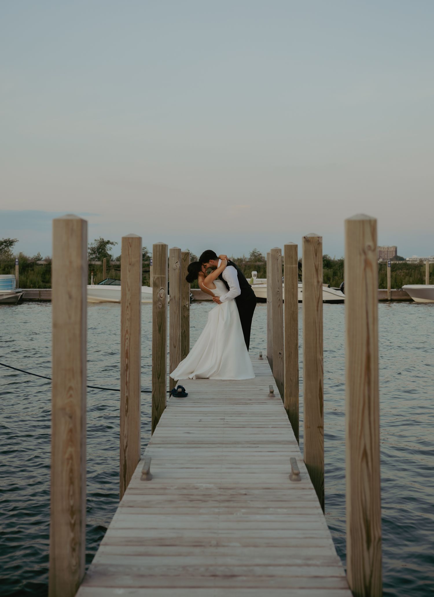 A bride and groom share a romantic kiss on a wooden dock over calm water at sunset during their wedding.