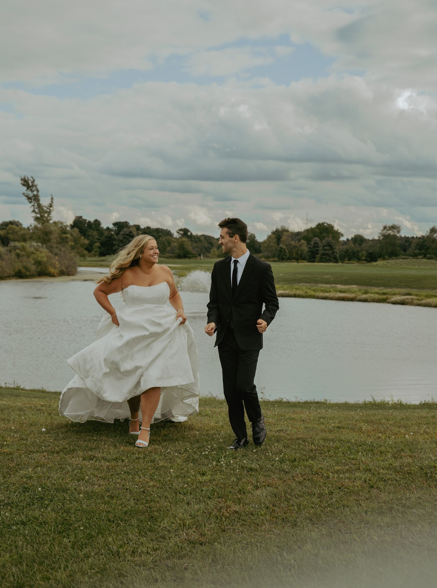 Bride in white gown and groom in black suit walk hand in hand beside a serene pond on their wedding day.