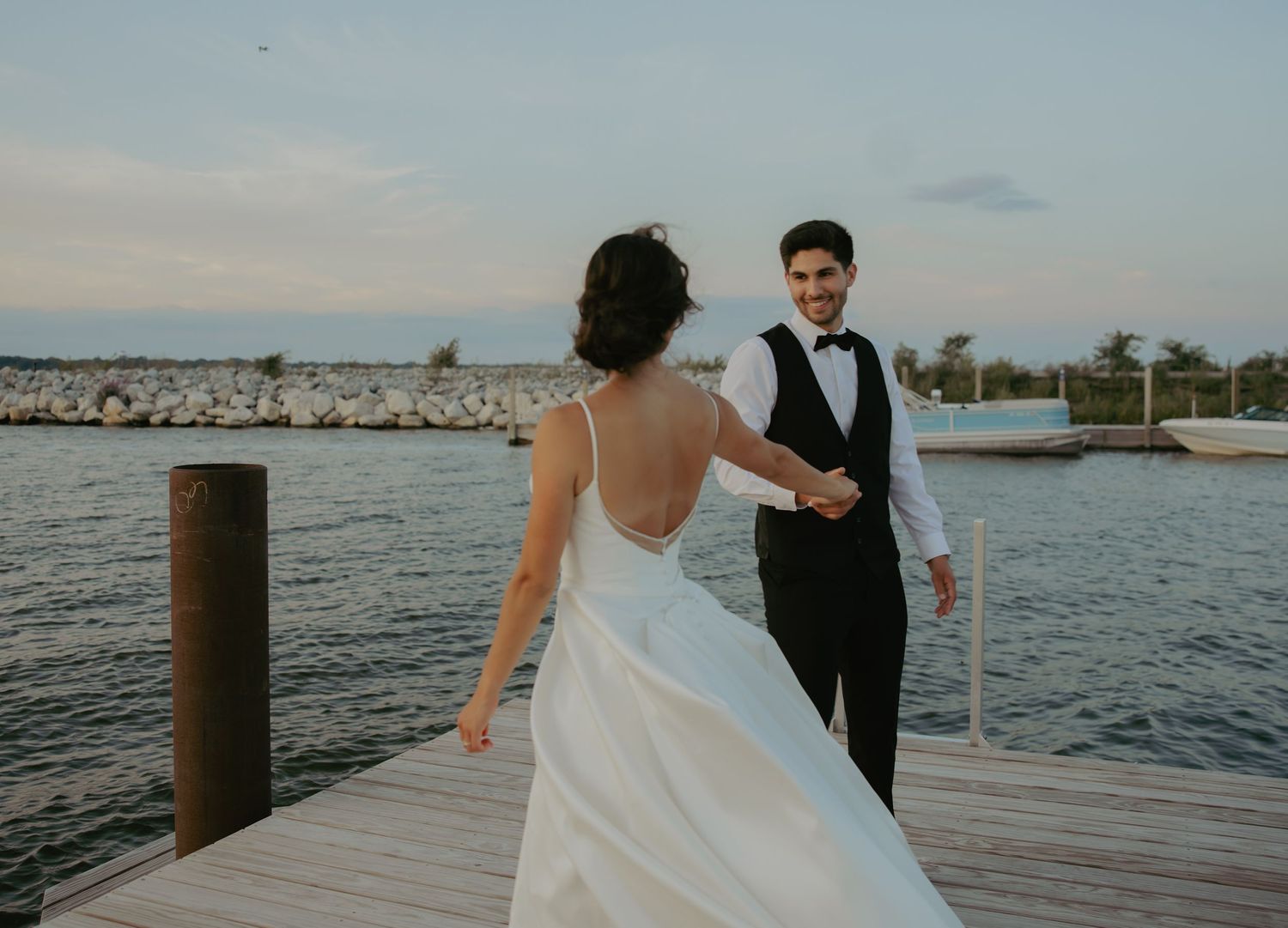 Bride and groom holding hands on a waterfront dock at sunset, bride in elegant backless white gown.