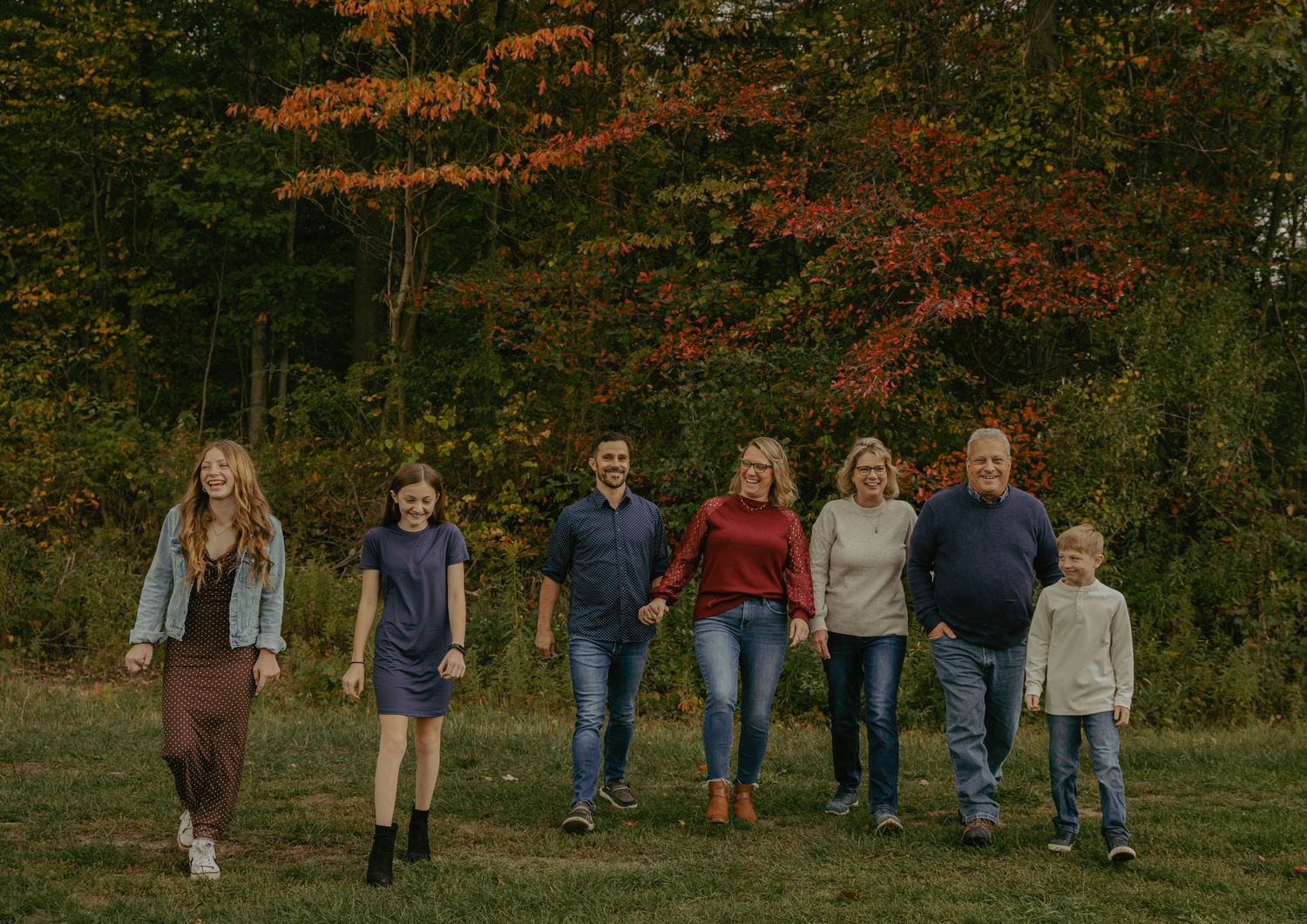 Family of seven walking together in a grassy field surrounded by colorful autumn foliage trees.