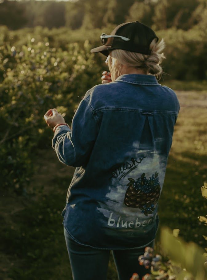 Back view of someone in a denim jacket with blueberry patch design picking berries.