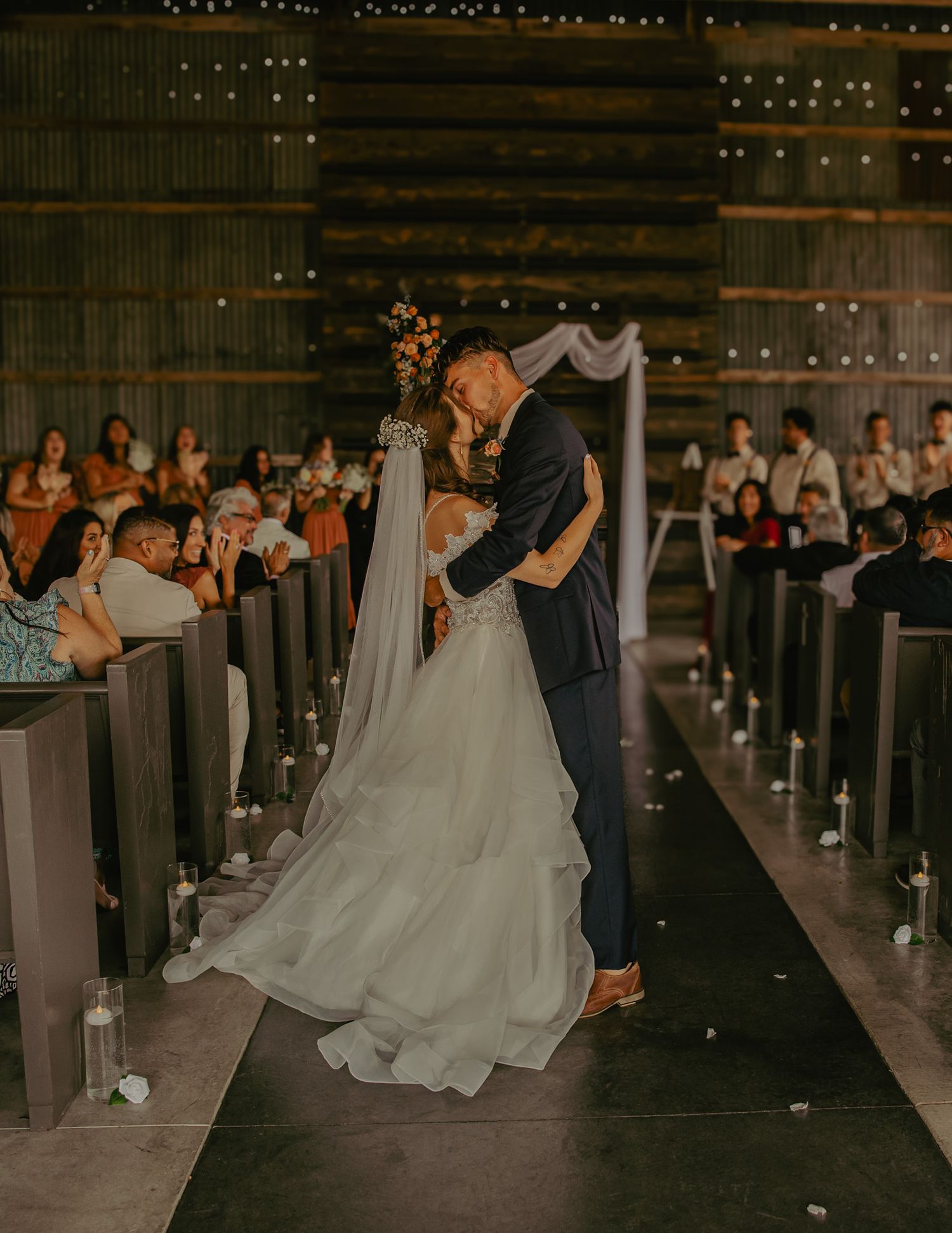 Wedding ceremony in rustic barn venue with string lights and draped aisle decorations.