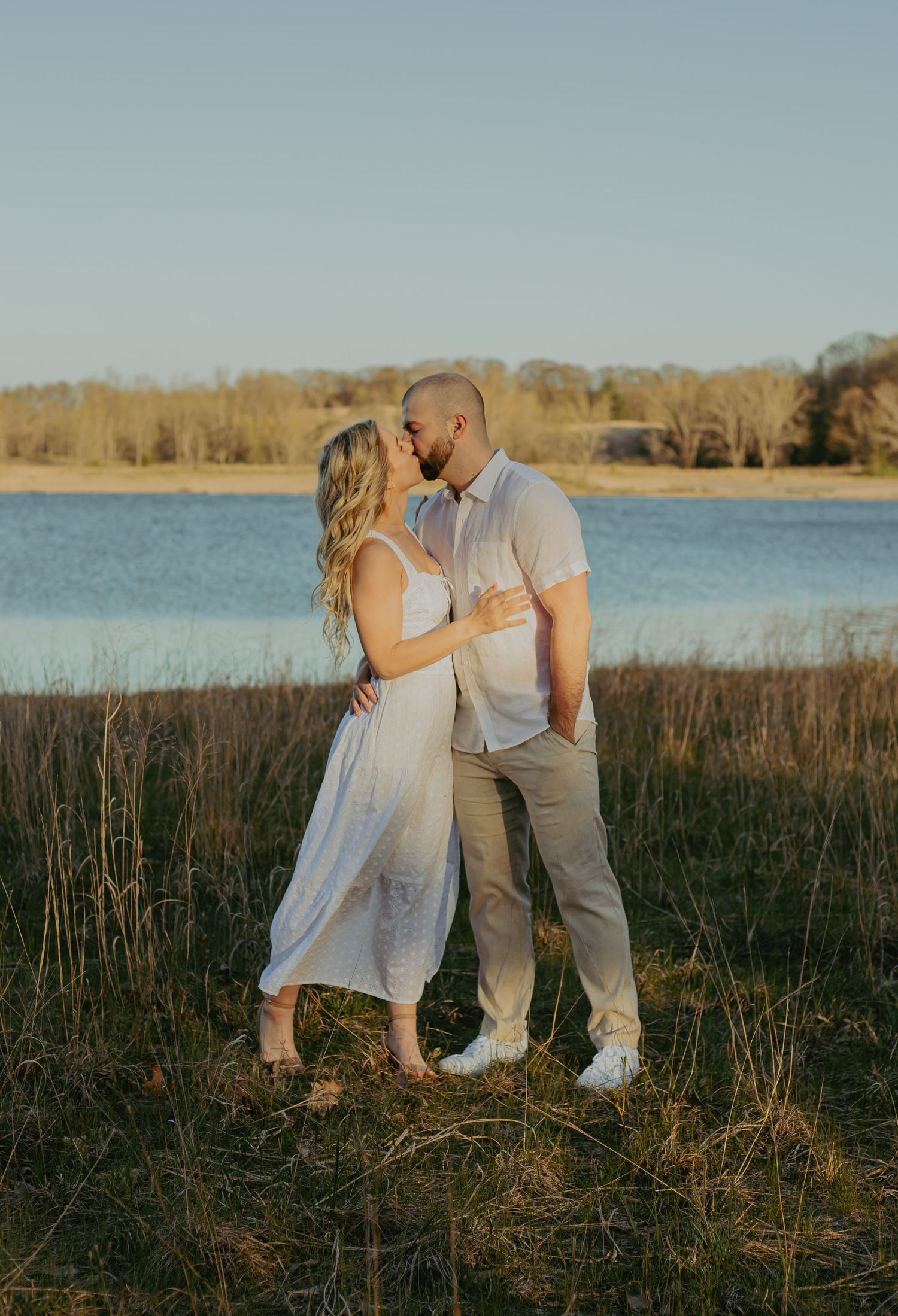 Couple in white outfits share intimate moment by lake with marsh grass at sunset.