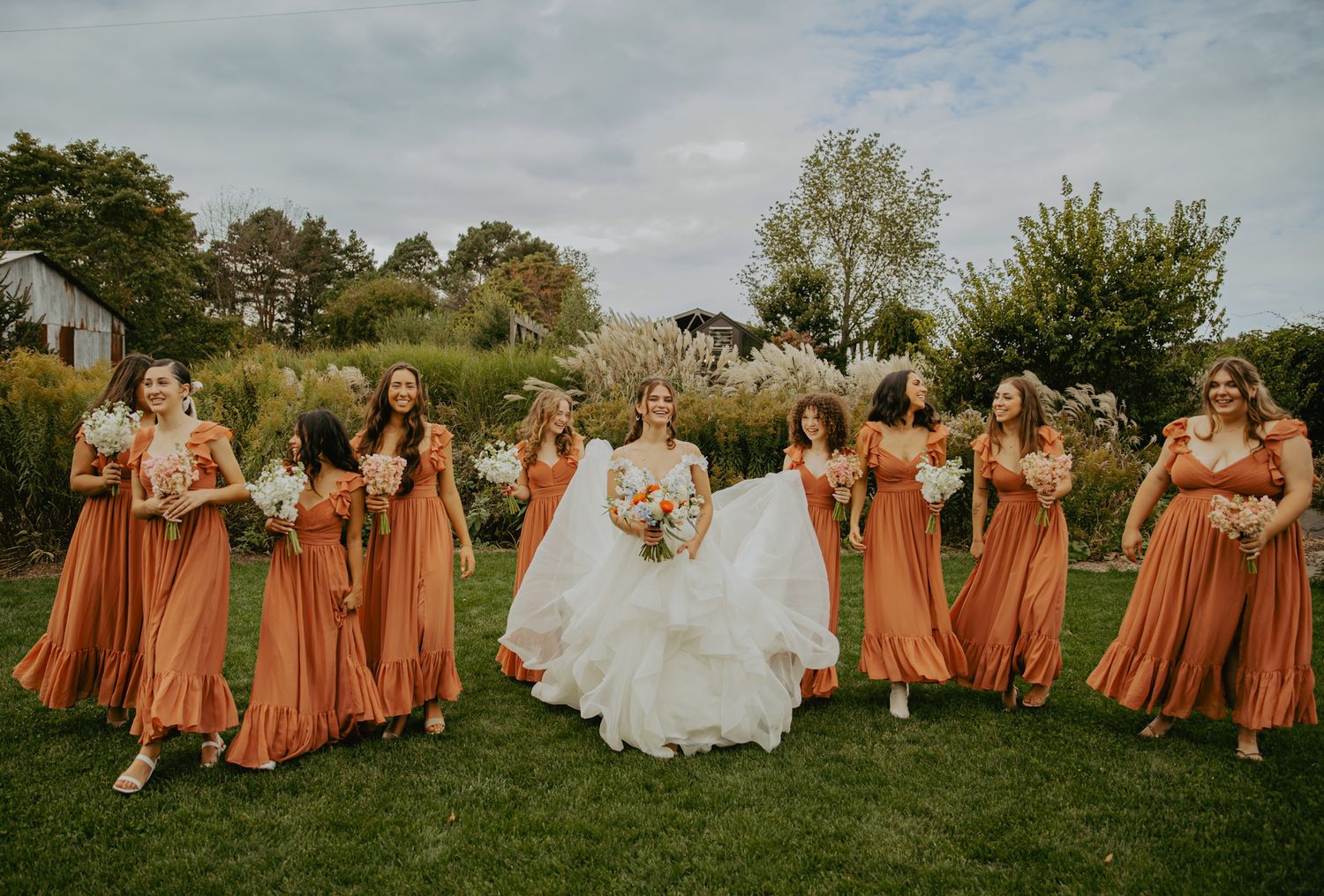 Wedding party in burnt orange bridesmaid dresses posing on grass under cloudy sky.