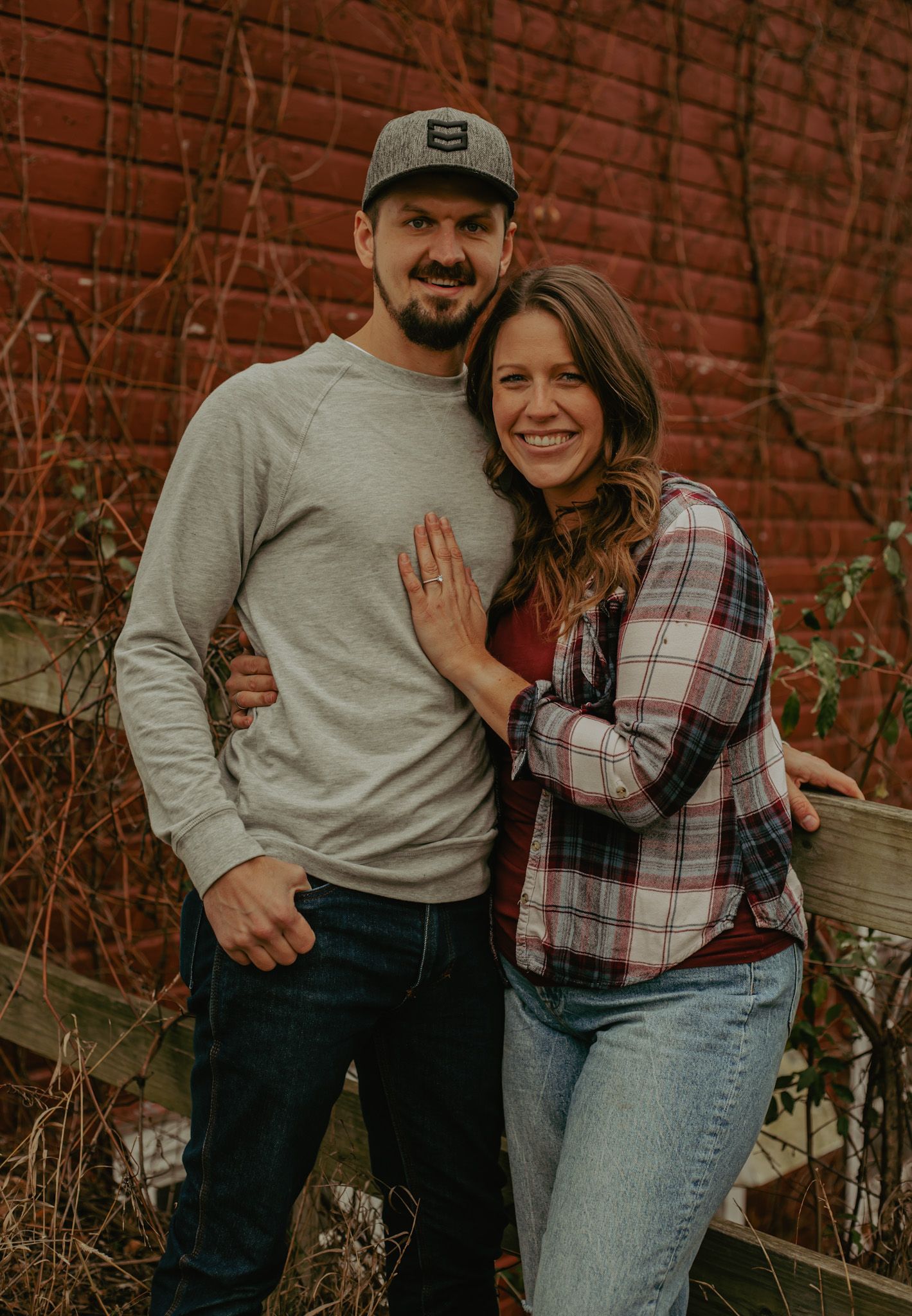 A couple poses casually against a red brick wall on an autumn day.